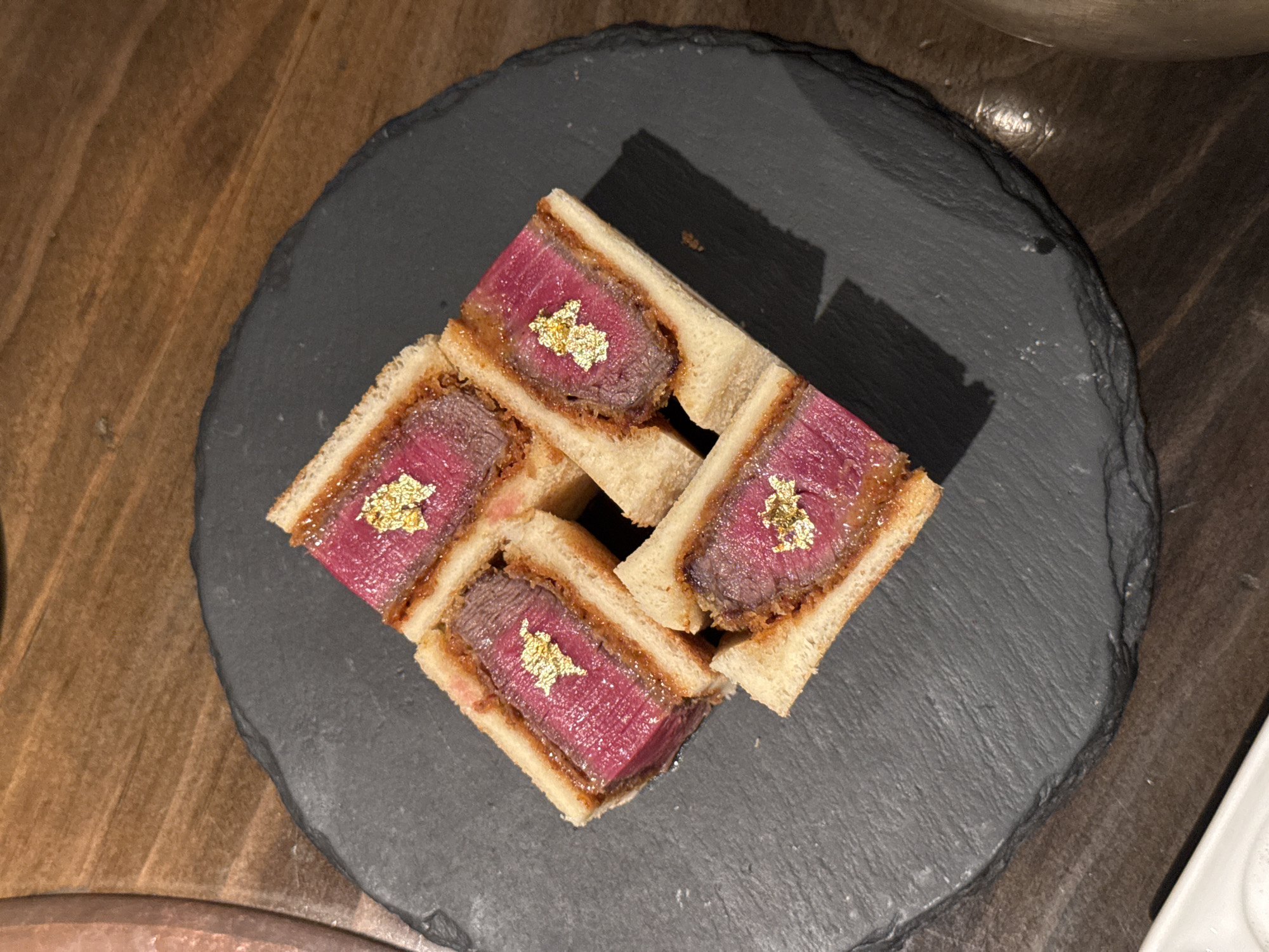 The image shows four square-cut sandwiches arranged on a round, dark gray slate serving plate. The sandwiches appear to contain a rare beef filling, each topped with edible gold leaf. The plate is set on a wooden surface. This is likely a food presentation in a restaurant setting, possibly in Seoul, South Korea. No people are visible.