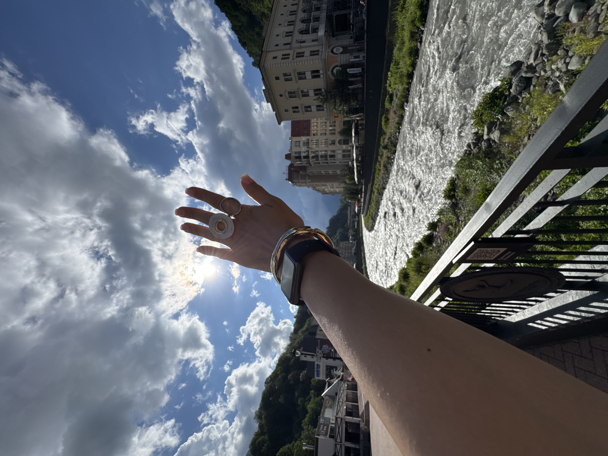 In the city of Estosadok, Russia, a person's arm is extended towards a bright sky filled with dramatic clouds and the sun. The person is wearing several rings on their fingers and a watch on their wrist. In the background, a river flows rapidly past a row of buildings, some of which appear to be hotels. Lush green hills rise behind the buildings, and trees are visible along the riverbank. The scene is captured on a sunny day with a mix of clouds and sunlight.