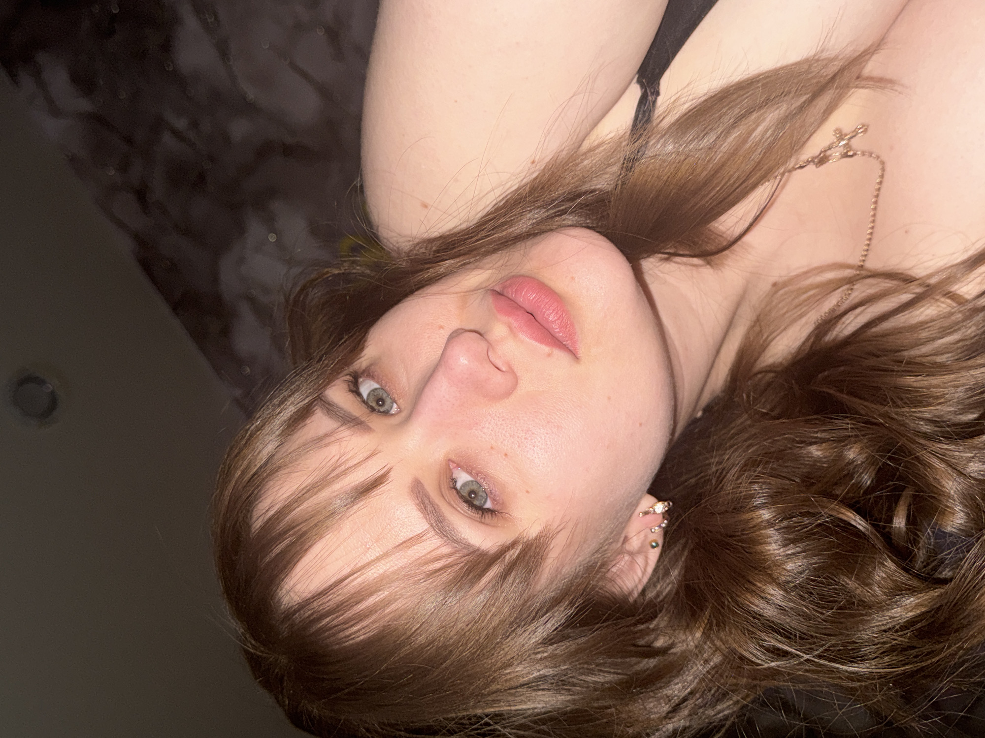 The image depicts a woman with light skin, brown hair, and green eyes. She is wearing a necklace with a cross pendant. She has multiple piercings in her ear. The background is a dark, blurry texture, possibly fabric or a wall. The image is taken at an unusual angle, with the woman's head tilted. The image location is not possible to identify from the context of the image.