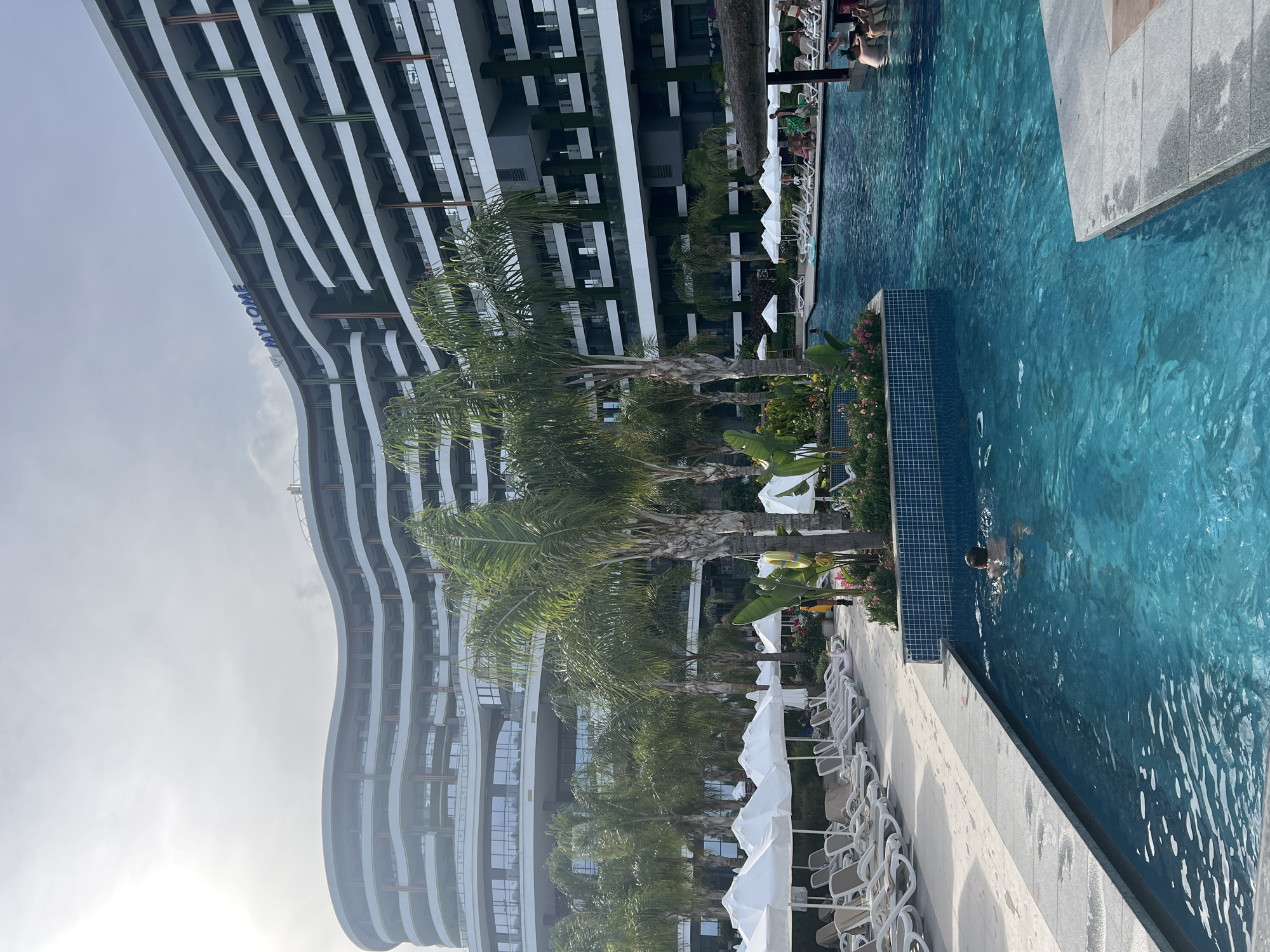 A man swims in a bright blue, outdoor swimming pool in front of a large, modern, curved hotel building. The hotel, with its white facade and multiple balconies, is visible in the background. Palm trees are scattered throughout the scene, adding a tropical feel. Beach chairs and umbrellas are lined up on the poolside, suggesting a resort setting. The sky is overcast, and the lighting indicates it's daytime. The hotel signage reads "MYLOME." This scene is consistent with resorts found in Alanya, Turkey, a popular tourist destination known for its beaches and hotels.