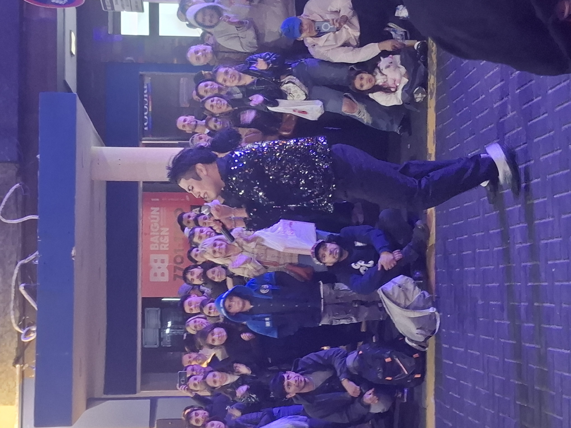 A crowd of people surrounds a performer wearing a sequined jacket and dark pants. The scene appears to be outdoors, on a brick-paved street in the city of Buenos Aires, Argentina. A blue awning and a sign with the name "Baigun" are visible in the background. Several people in the crowd are holding up phones, possibly recording or taking pictures of the performer.