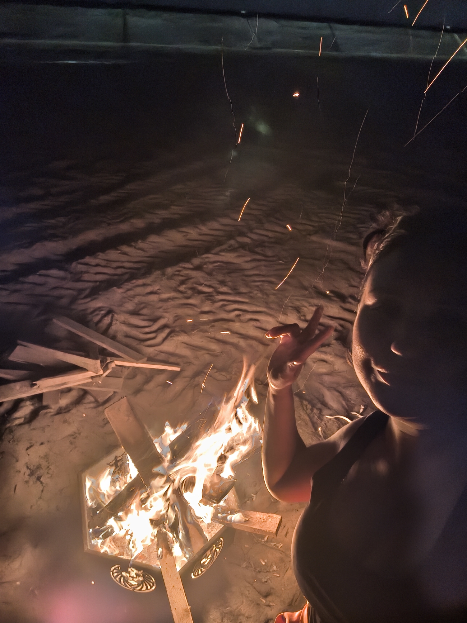 A high-angle, medium-shot photograph taken at night, appears to depict a person standing next to a fire pit in Сокуры, Russia. The person is partially visible on the right side of the frame, with their face and torso illuminated by the light of the fire. They are gesturing with their left hand. In front of the person is a square fire pit, filled with burning wood and flames. The fire throws light and sparks upwards, some of which are captured in the air around the person. The background is a dark, textured surface that appears to be a blanket or tarp spread out on the ground. Additional pieces of wood are scattered around the fire pit.