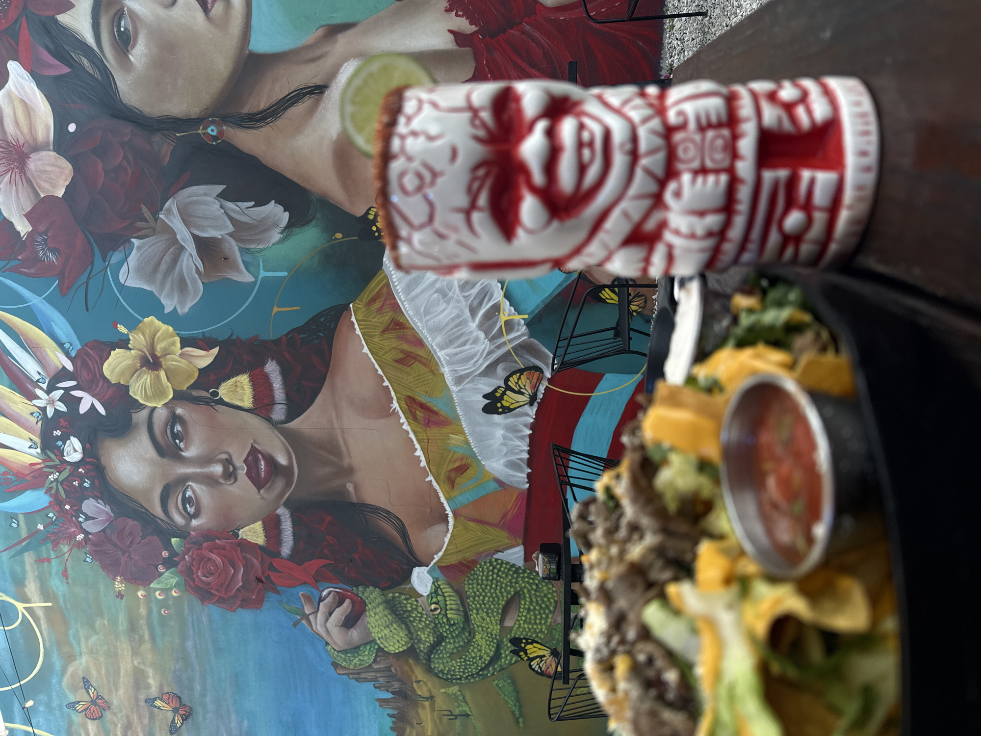 The image presents a vibrant and art-filled dining scene, likely from a restaurant or bar in Hollywood, United States of America. The primary backdrop is a large, colorful mural featuring two stylized female figures.
The upper figure in the mural is partially visible, showing dark hair, a subtle eye, and a bare shoulder adorned with large white and red flowers. A lime slice, presumably from a drink not fully in frame, is positioned near her face. The more prominent lower figure is depicted upside down relative to the viewer's orientation. She wears an elaborate headdress of yellow, red, and white flowers, feathers, and several small blue butterflies. Her face shows a calm or pensive expression, with dark eyes and reddish lips. She is wearing a vibrantly patterned yellow and white garment. Her visible arm holds a red fruit, possibly an apple, around which a green snake is coiled. Additional yellow and orange butterflies are scattered across the mural, which features swirling patterns in blue, teal, and gold.
In the foreground, on a dark wooden table, is a distinctive white and red tiki mug. It is elaborately decorated with stylized red patterns and faces, suggesting a tropical or themed cocktail. A lime wedge is visible, indicating the drink within. Adjacent to the mug is a dish of food, partially out of focus, which appears to be nachos. The dish contains tortilla chips, ground meat, shredded lettuce, and melted cheese, with a small black cup of red salsa or dip placed in the center.
The overall scene conveys a casual and visually rich dining experience, likely during the day or evening, in a uniquely decorated establishment. No discernible text is visible.