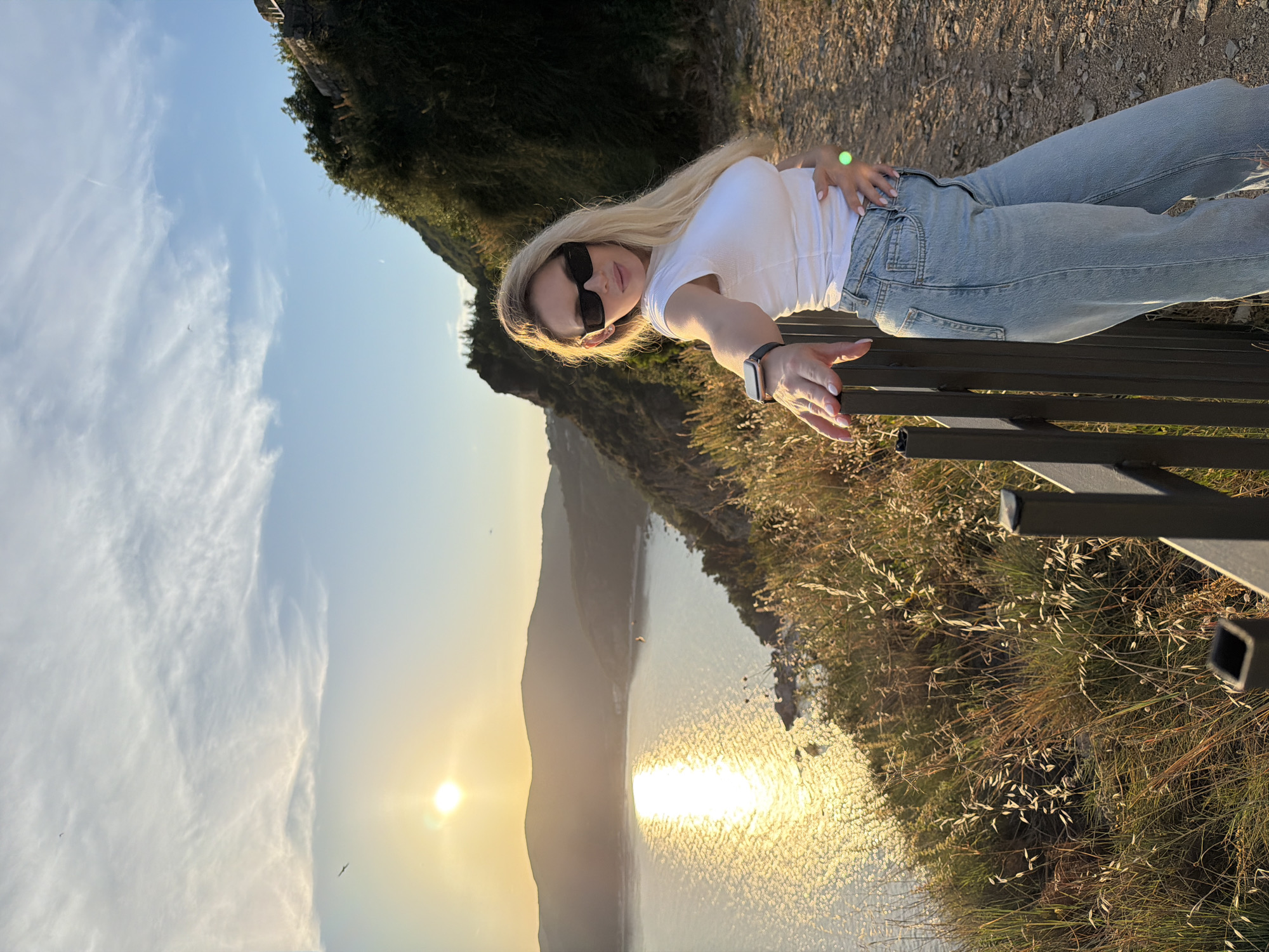 A woman with blonde hair, wearing a white t-shirt, blue jeans, and black sunglasses, poses on a path overlooking a body of water at sunset. The sun, low on the horizon, casts a golden glow on the water, creating a shimmering effect. Mountains rise in the background, covered in lush green foliage. The woman is leaning against a dark metal railing, her right hand resting on it, while her left hand is placed on her stomach. A smartwatch is visible on her left wrist, and a green ring is on her right ring finger. The path is made of gravel, and dry grass and some small plants grow along the edge of the path and the railing. In the distance, a few birds can be seen flying in the sky. The scene appears to be located on a cliffside overlooking a bay, likely within the vicinity of Budva, Montenegro, given the characteristic coastal landscape.