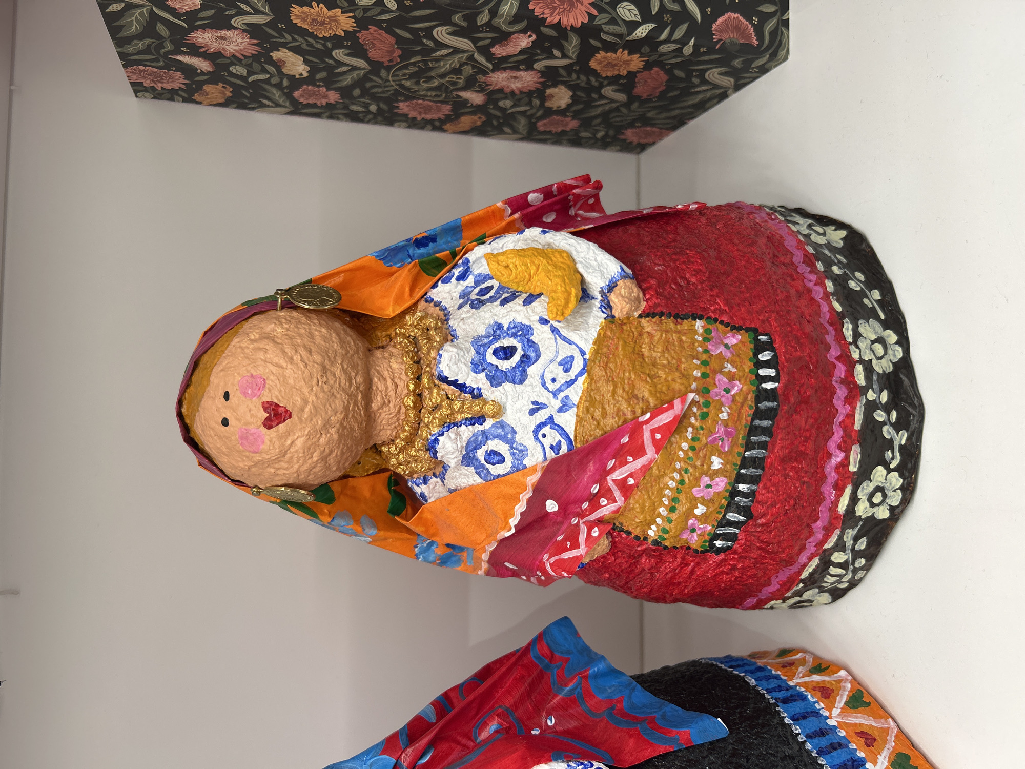 The image shows a close-up, eye-level view of a handmade doll, likely made from paper-mâché, displayed on a white shelf. The doll is adorned in traditional-looking clothing with vibrant colors and intricate patterns. It wears a red skirt with a black and white floral border, a white blouse decorated with blue floral motifs and small birds, and an orange and pink patterned headscarf with gold trim. The doll has a textured, light brown face with painted pink cheeks, black eyes, and a red heart-shaped mouth. It also wears a gold necklace and a gold earring on its right side. A small, yellow, boot-shaped object is held in the doll's right hand. To the left of the doll, a portion of another similar doll is visible, wearing a red and blue garment. To the right, a dark-colored decorative box with a floral pattern is placed. The background consists of a plain white wall and shelf. The lighting appears to be even and diffused, suggesting an indoor setting.