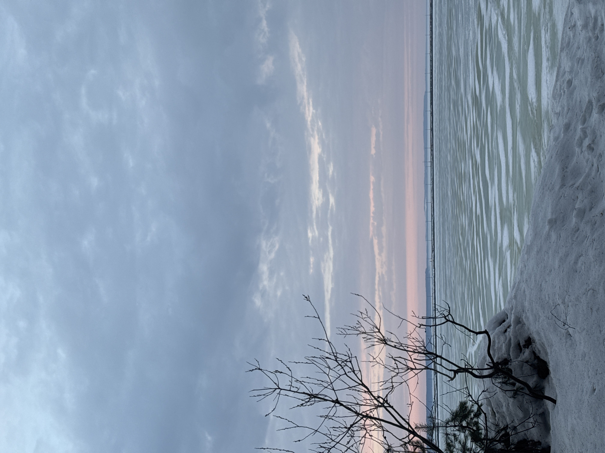 The image shows a winter scene in Chaikovsky, Russia. In the foreground, bare branches of a tree reach into the sky. Patches of snow cover the ground. Beyond the snow and branches, a frozen body of water stretches to the horizon. Wind turbines are visible in the distance. The sky is overcast with streaks of pink along the horizon.