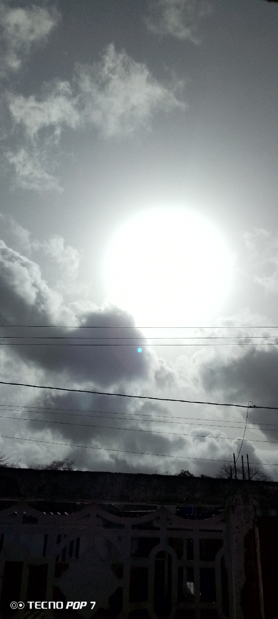 The image depicts a bright sun partially obscured by clouds, creating a high contrast scene. Several power lines are visible in the midground, running horizontally across the frame. Below the power lines is a dark, solid structure, possibly a rooftop. In the foreground is a pale concrete fence or railing, partially obscuring the view. The image was taken with a Tecno Pop 7 smartphone. Based on the prompt, the location is Altagracia, Venezuela.