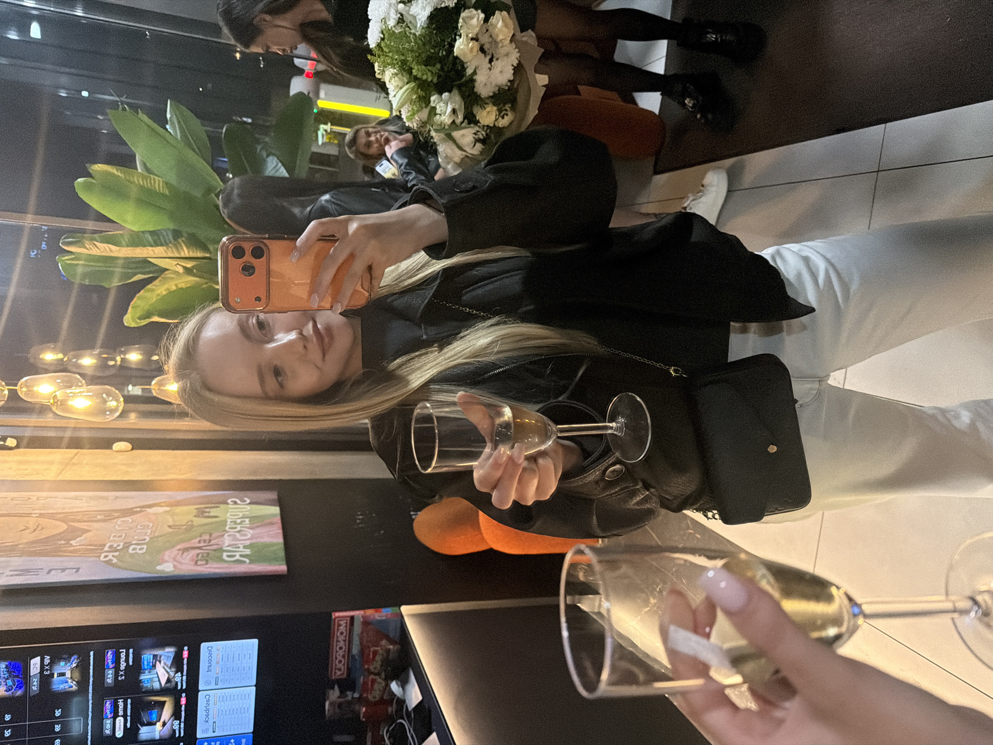 Here is a detailed description of the image:
The image depicts a scene from what appears to be a social gathering inside a venue with modern interior design. A blonde woman is prominently featured, holding a champagne flute in one hand and using her orange smartphone to take a photo or video with the other. She is wearing a black leather jacket, white pants, and a black purse hanging on her shoulder.
To the right of the woman is another champagne flute held by a hand with manicured nails, suggesting another person is present. In the background, there are other individuals, one holding a bouquet of white flowers. A plant is also visible.
The setting includes a TV screen displaying various content, a piece of art on the wall above the TV, and modern lighting fixtures.