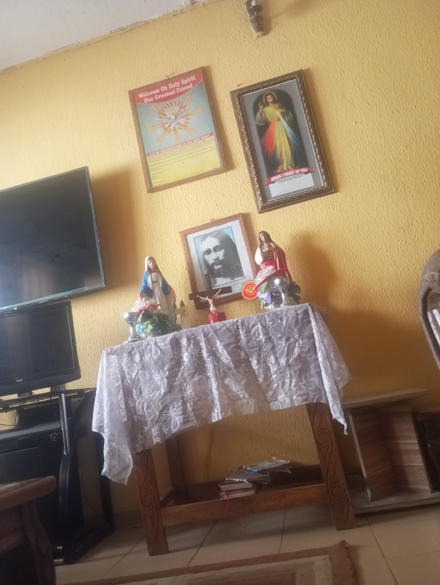 The image captures a scene in a room, possibly in Onitsha, Nigeria, characterized by religious iconography and furnishings. The dominant wall color is a textured yellow. On the wall, two framed religious prints are visible. The print on the left is titled "Welcome Oh Holy Spirit Our Greatest Friend" and depicts a dove surrounded by radiant lines. The print on the right shows an image of Jesus with the text "JESUS I TRUST IN YOU" at the bottom.
In the foreground, a wooden table is draped with a white lace tablecloth. On the table, there are religious figurines: a statue of the Virgin Mary, a statue of Jesus on a crucifix, and another statue of Jesus with his hand raised. A silver chalice and some artificial flowers are also present. Behind the table, a dark-colored television set is visible, mounted on a stand. To the left of the table, another, smaller television or monitor is positioned on a stand.
The overall impression is that of a domestic or devotional space, likely within a home. The lighting suggests an indoor setting, possibly with natural light filtering in from an unseen source. The floor appears to be tiled. The presence of multiple religious items indicates a strong religious devotion within the household.