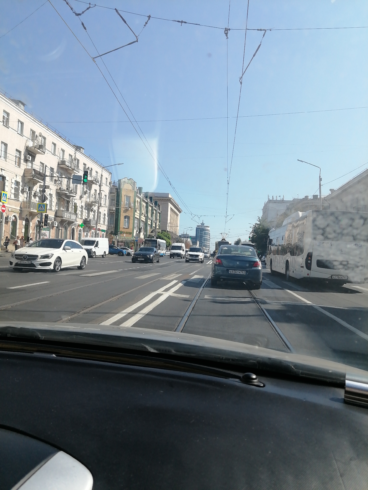 The image shows a street scene in Rostov-on-Don, Russia, likely on a sunny day. The view is from inside a vehicle, looking forward through the windshield. In the foreground, the dashboard of the vehicle is visible. The street is paved and has tram tracks. Several cars, including a white Mercedes, a dark sedan, and a white van, are driving on the road. A large white bus is also visible. In the background, there are multi-story buildings with balconies, some with air conditioning units. There are also traffic lights, streetlights, and overhead wires, indicating a well-developed urban environment. Pedestrians can be seen walking on the sidewalk near the buildings. The sky is clear and blue. The overall impression is of a typical city traffic scene.