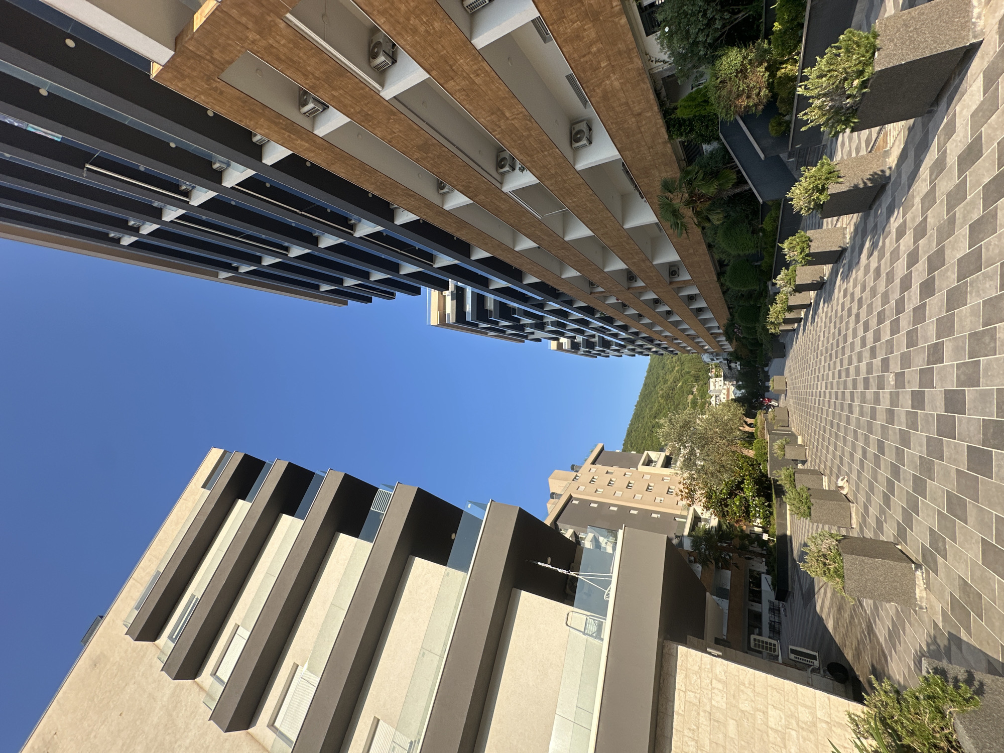 The image depicts a contemporary street scene in Budva, Montenegro, characterized by modern multi-story buildings lining a pedestrian walkway. The walkway is paved with a repeating pattern of light and dark gray square tiles, laid diagonally to the direction of the path. On either side of the walkway, large, angular planters contain green, shrub-like vegetation.
To the left, a light-colored building with beige and dark gray balconies dominates the foreground. The building features prominent horizontal dark gray beams that extend outwards, supporting balconies with glass railings. Windows with light-colored blinds are visible behind the railings.
To the right, a taller building with a more extensive facade of alternating light and dark horizontal elements rises into the sky. This building has numerous balconies, each with a white inner section and a dark gray exterior. Many of these balconies have visible outdoor air conditioning units mounted on their underside. The facade of this building also features sections with a warm, brown, wood-like texture.
In the distance, a lighter colored, more traditionally styled building with multiple windows is visible, situated between the two modern structures. Further back, a lush green hillside with some scattered buildings and trees forms the backdrop. The sky is a clear, vibrant blue, suggesting a sunny day. A single person, appearing as a small, dark figure, is walking down the central pathway in the distance. A dog is also visible lying down on the walkway near one of the planters.