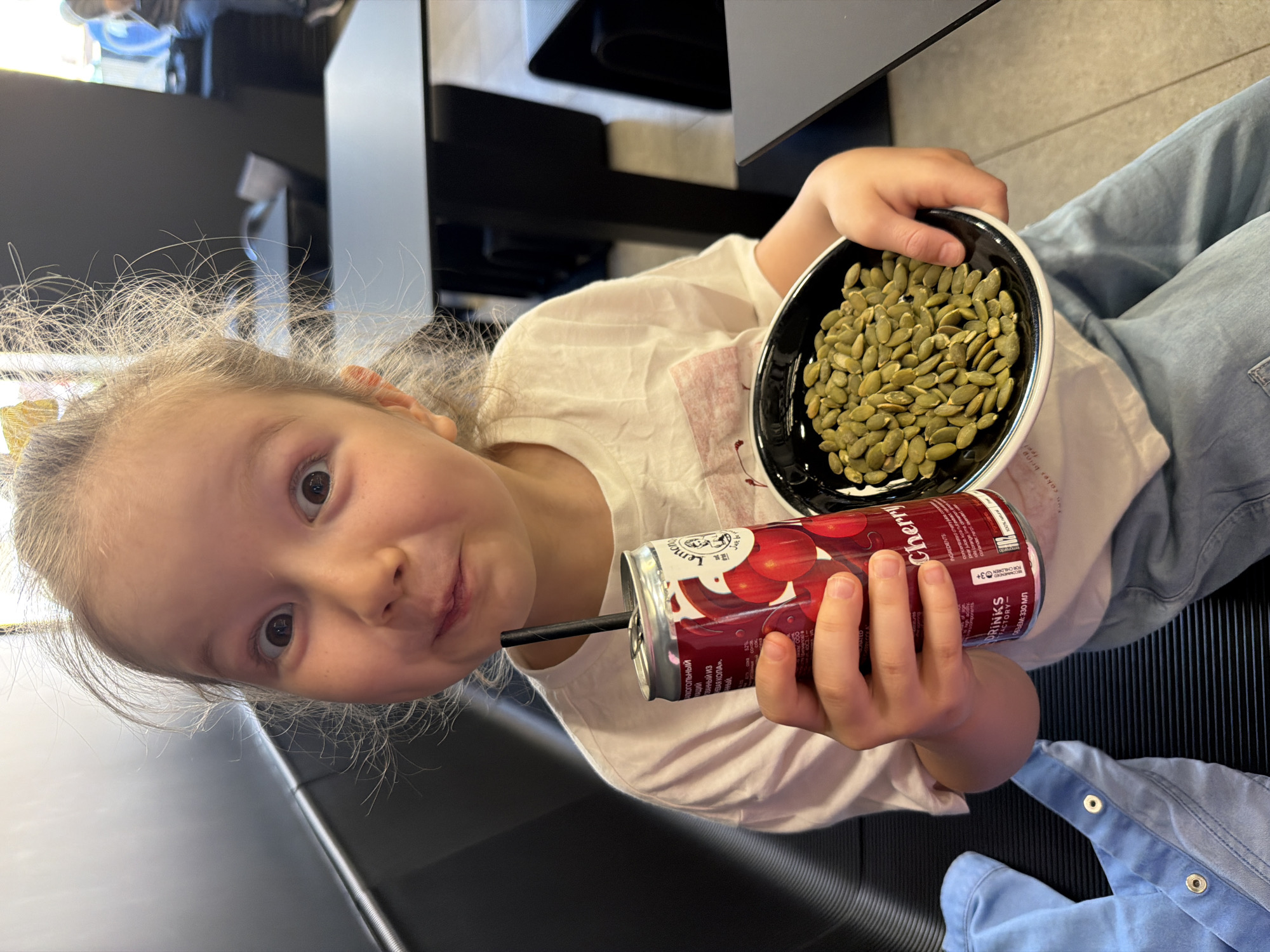 A young girl with fair hair, wearing a white t-shirt and blue jeans, sits at a table and holds a can of "Cherry" Lemon drink in her right hand, with a black straw in the opening, and a black bowl filled with green pumpkin seeds in her left hand. The can's label includes text in Russian, "АЛКОГОЛЬНЫЙ ВАННЫЙ ИЗ ВАЯ КОЛА." and " Lemon". The label also features the text "FOR CHILDREN 3+". A portion of a blue shirt is visible in the lower right corner. The table has a black surface, and background elements include furniture and a wall. The location is Izhevsk, Russia.