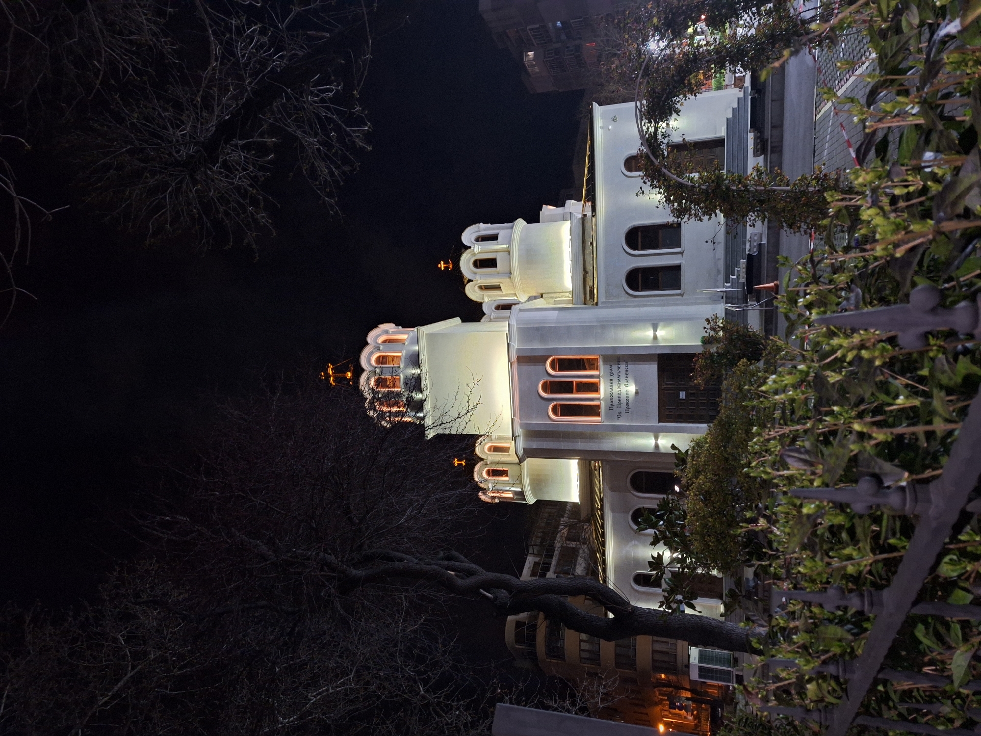 The image shows the Orthodox Cathedral of the Dormition of the Mother of God in Varna, Bulgaria, at night. The white facade of the building is illuminated, highlighting its architectural details, including arches and towers. The cathedral is partially obscured by trees and bushes, and a wrought iron fence runs along the foreground. The background is dark, with a glimpse of buildings illuminated by warm lights.