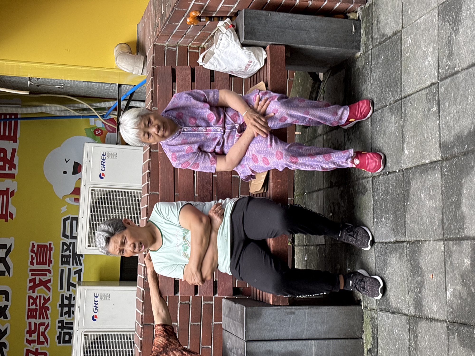 Two elderly Asian women are sitting on a slatted wooden bench in an outdoor urban setting. The woman on the left has short, graying hair and is wearing a light green t-shirt and black pants. Her arms are crossed in front of her chest, and she appears to be speaking. The woman on the right has short, white hair and is wearing a purple pajama-like set with a floral pattern. She is looking forward, with her hands resting on her lap. Behind them, there are two white air conditioning units mounted on a wall, with Chinese text and graphics on a yellow background above them. To the right of the bench, there is a white reusable shopping bag and an orange object, possibly a cane or part of a building fixture, attached to the brick wall. The ground is paved with gray square tiles, and to the left of the bench, a dark gray concrete structure is visible. The overall environment suggests a commercial or residential area in Yuzhong District, China.