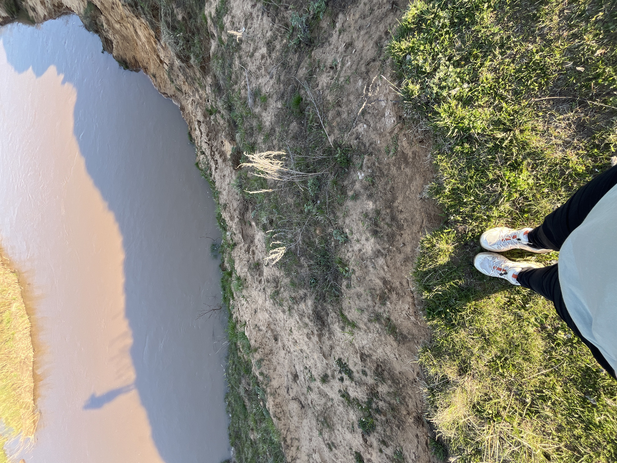A high-angle shot depicts a person standing on the edge of a cliff overlooking a body of water, likely a river. The person is wearing white running shoes, black leggings, and a light blue shirt. The cliff edge is composed of dirt and sparse vegetation. The water appears muddy, with a light brown hue on one side, potentially indicating a different water source or sediment concentration. The opposite bank has green vegetation. The scene suggests an outdoor location with a natural feature like a river valley or ravine. It is impossible to pinpoint Воронино, Russia as the location as no landmarks are apparent.