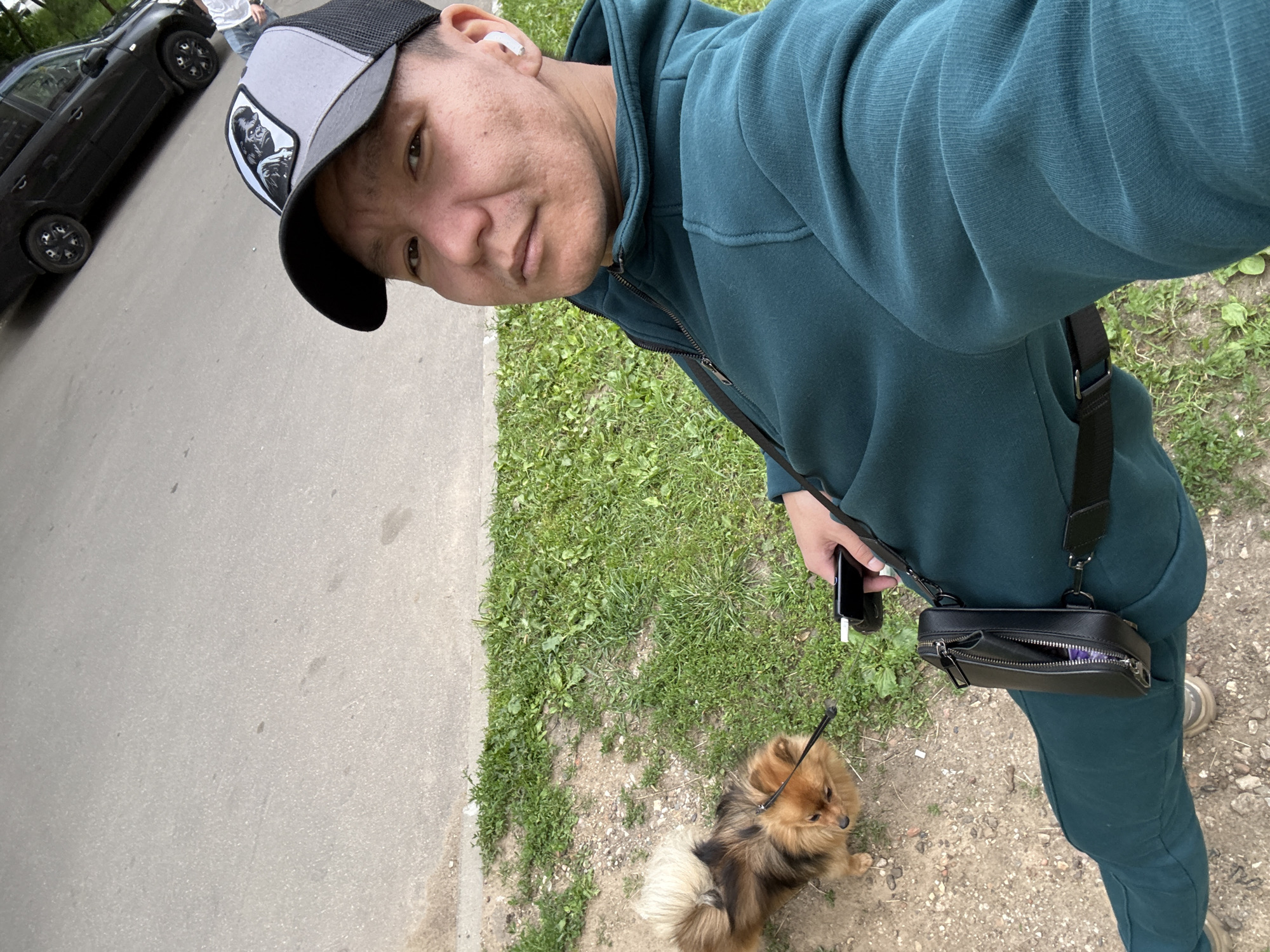 The media file shows a person, likely male, taking a selfie outdoors. The person is wearing a teal hooded sweatshirt and matching pants, a black and grey baseball cap with a graphic on the front, and earbuds. They are holding a smartphone in their right hand. To their left, a small, fluffy brown dog on a leash is visible, looking to the right. The background includes a paved area, a grassy patch with some dirt, and a black car parked in the distance. The image appears to have been taken during the daytime, with natural light illuminating the scene. The overall mood seems casual and relaxed. There is no visible text in the image. The context provided suggests the location is Moscow, Russia.