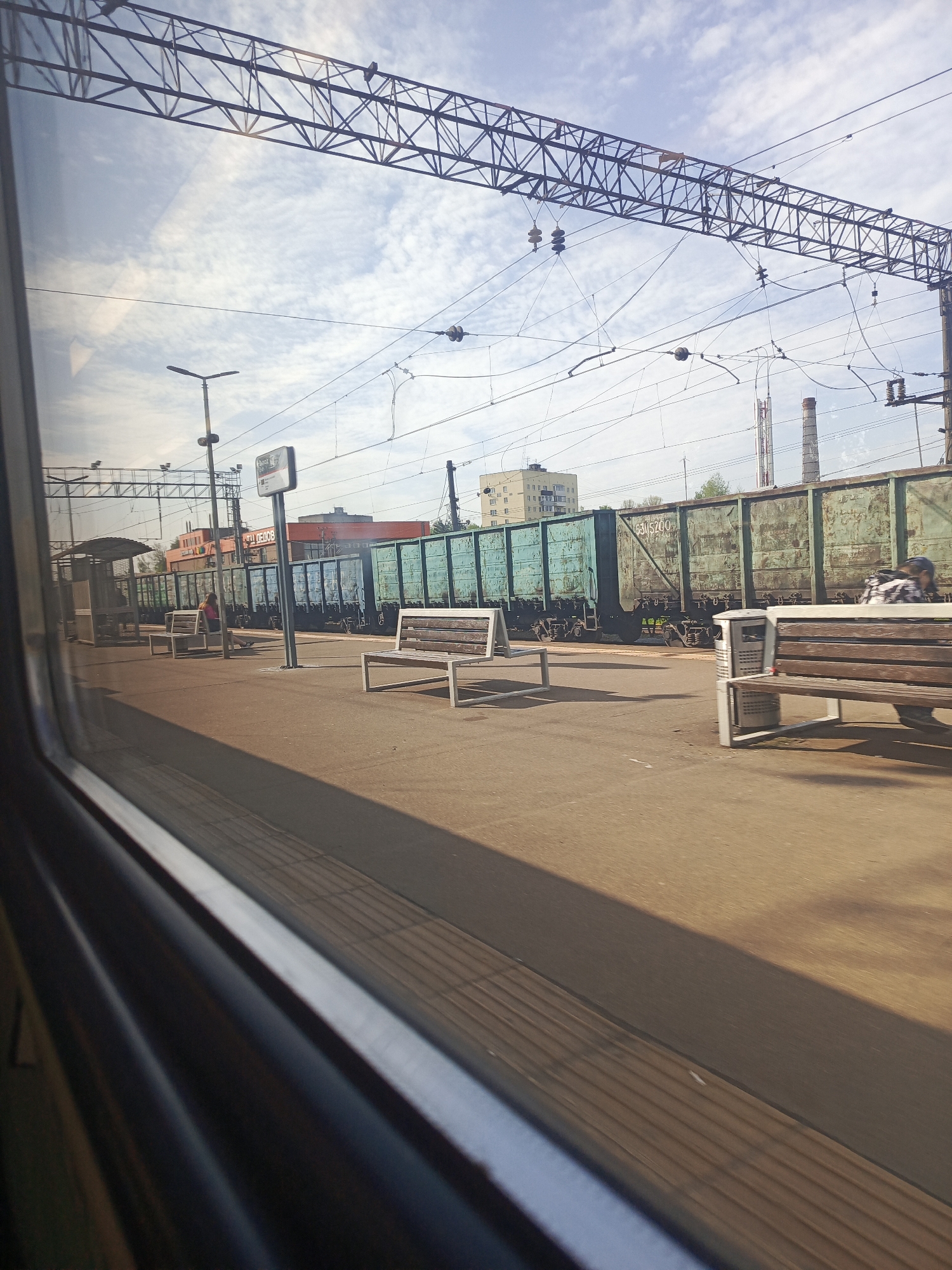 The image, taken from the interior of a moving train, shows a train station platform in Dedovsk, Russia. The platform is made of concrete and has several wooden benches with white metal frames. People are visible on the platform, either sitting on the benches or walking. Behind the platform, there is a freight train composed of green train cars, parked on the tracks. In the background, there are buildings of varying sizes, including what appears to be a residential building and industrial chimneys. Overhead, there is a network of power lines and metal structures associated with the railway infrastructure. The sky is cloudy. The lighting is bright, indicating it is daytime. The view is partially obstructed by the train window's frame, which introduces a dark, blurry foreground.