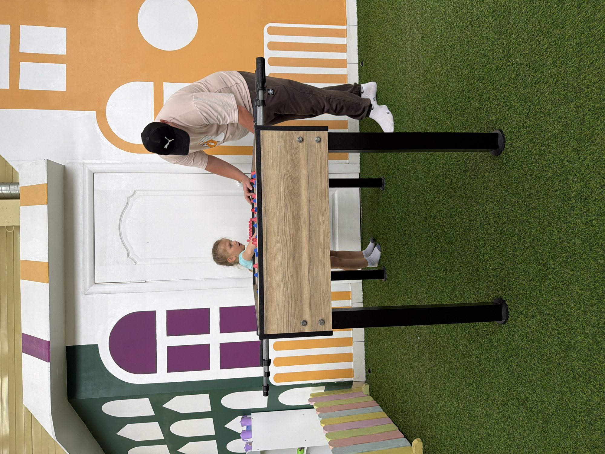 A medium shot captures a man and a young girl playing foosball in a brightly colored indoor play area. The man, wearing a black baseball cap, beige t-shirt, and brown pants, is bent over the table, his hand on the foosball handle. The girl, wearing a blue shirt, is looking up at him from the other side of the table. The foosball table is wood-grain and black, set against a backdrop of faux grass and a wall painted with a cartoonish house design in various colors including orange, white, purple, and green. The scene is likely in a children's play center, possibly in the city of Chaikovsky, Russia.