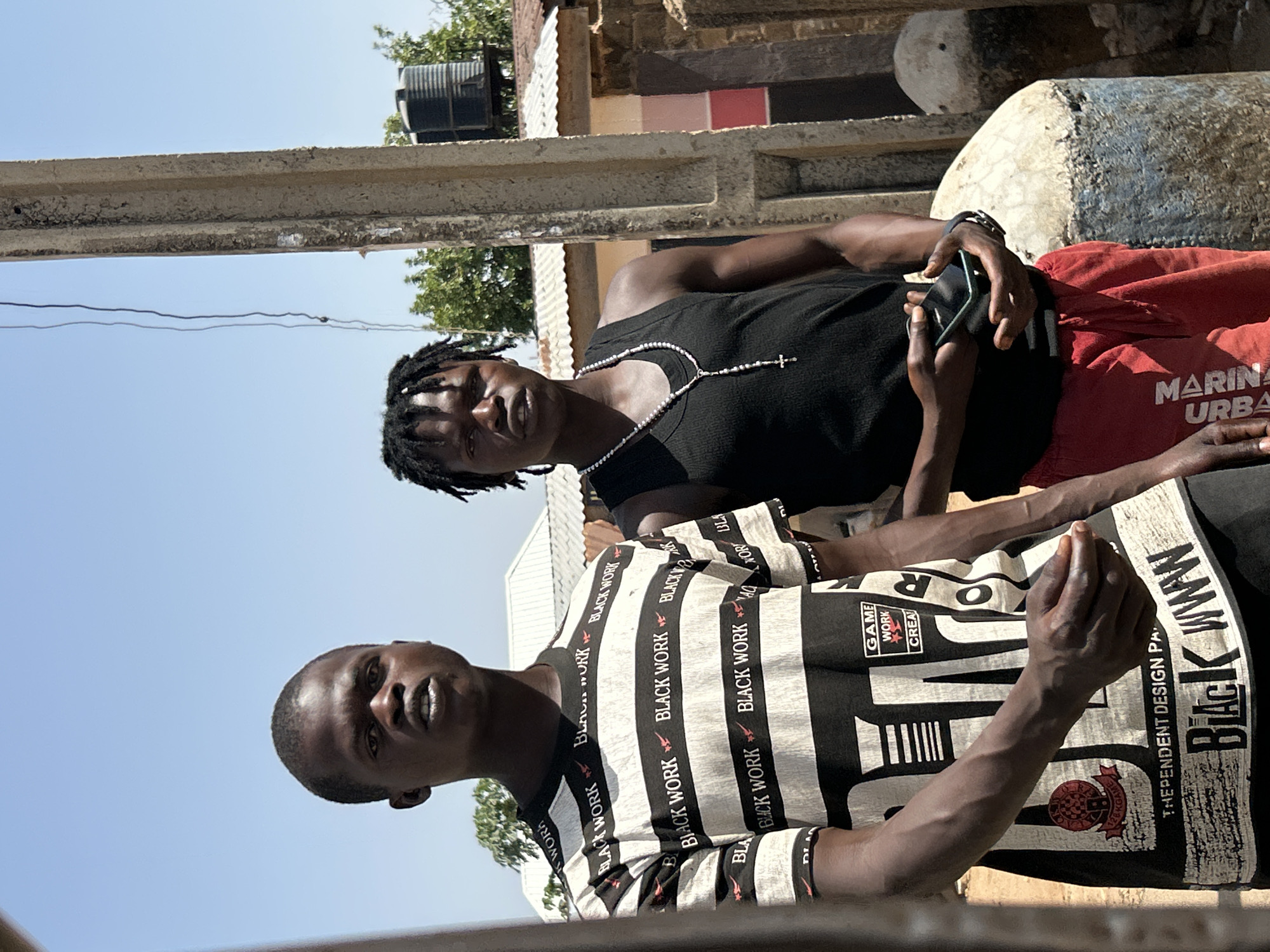 Two men are sitting outside. One is wearing a black tank top with a beaded necklace and red shorts. The other man is wearing a black and white striped shirt that says "BLACK WORK". The location is most likely in the city of Jos, Nigeria.