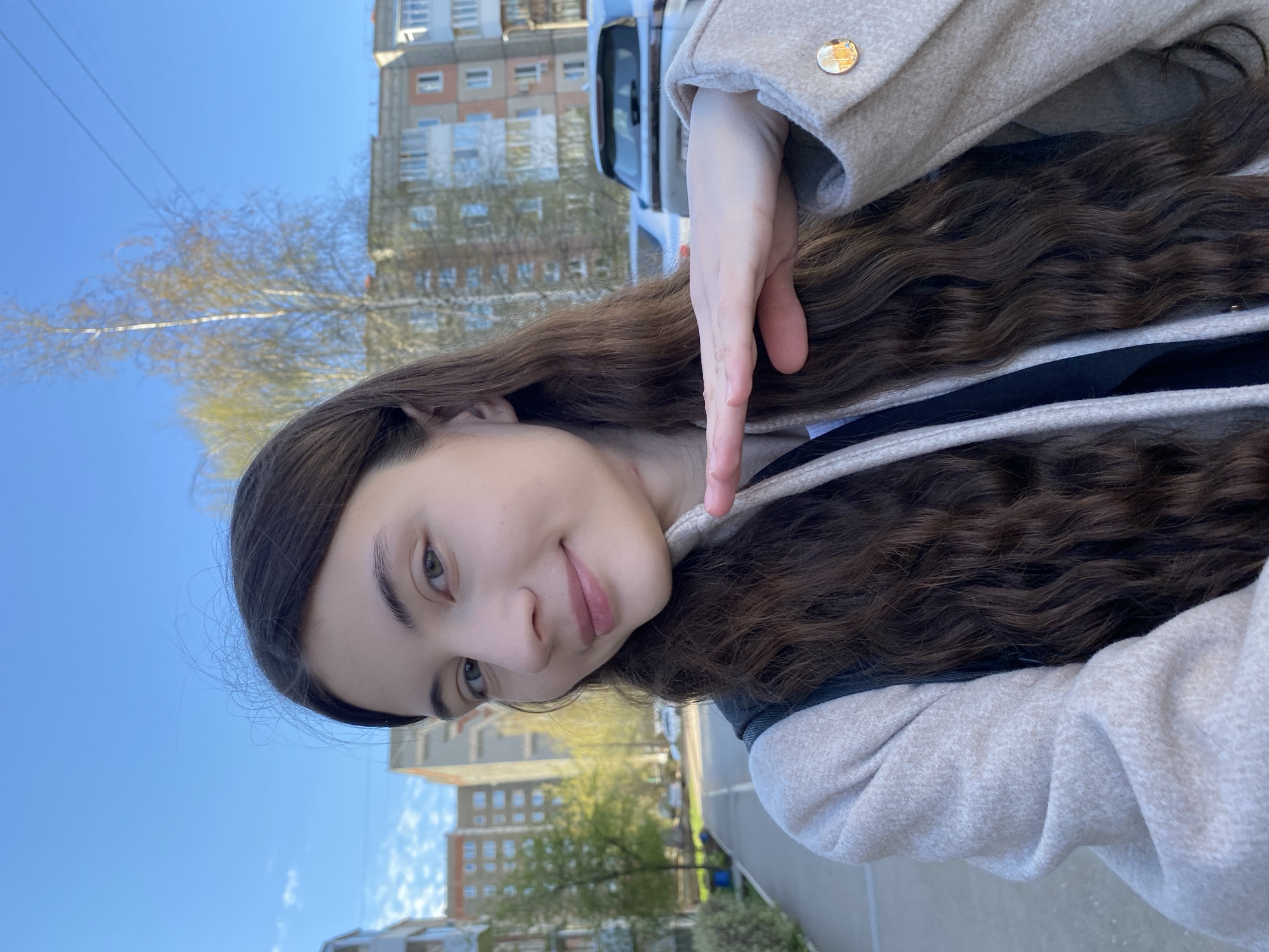 A woman with long, dark wavy hair poses for a selfie outdoors. She wears a light-colored coat with a gold button on the sleeve. Her hand is raised near her face. Behind her, there's a multi-story building, a tree with budding leaves, and a blue sky. Based on the architectural style and vegetation, the location appears to be in a residential area, likely in Votkinsk, Russia. A car is visible behind her.