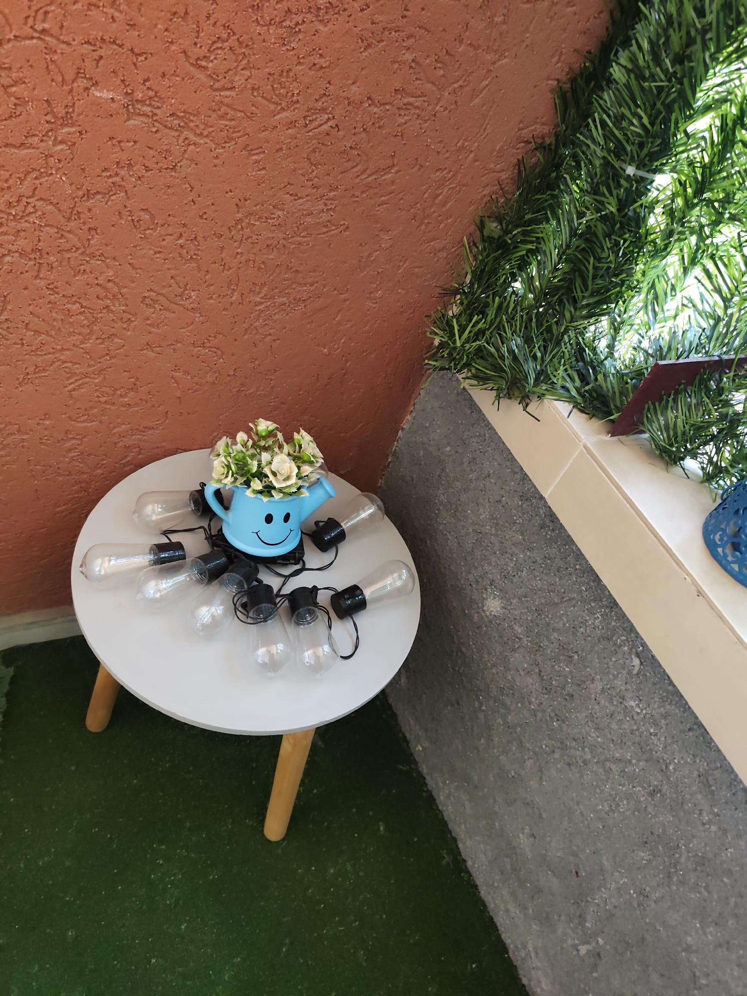 A medium shot shows a small round white table with wooden legs positioned in the corner of a space, likely a patio or balcony. The table is topped with a string of lights arranged around a small, blue watering can with a smiling face, which is filled with small white and green flowers. The table is sitting on a surface that appears to be artificial green grass.
Behind the table is a wall with textured orange paint. To the right of the table, there is a low wall with a rough grey surface and a tiled top, which is decorated with artificial green garland. The scene is probably located in Plovdiv, Bulgaria.