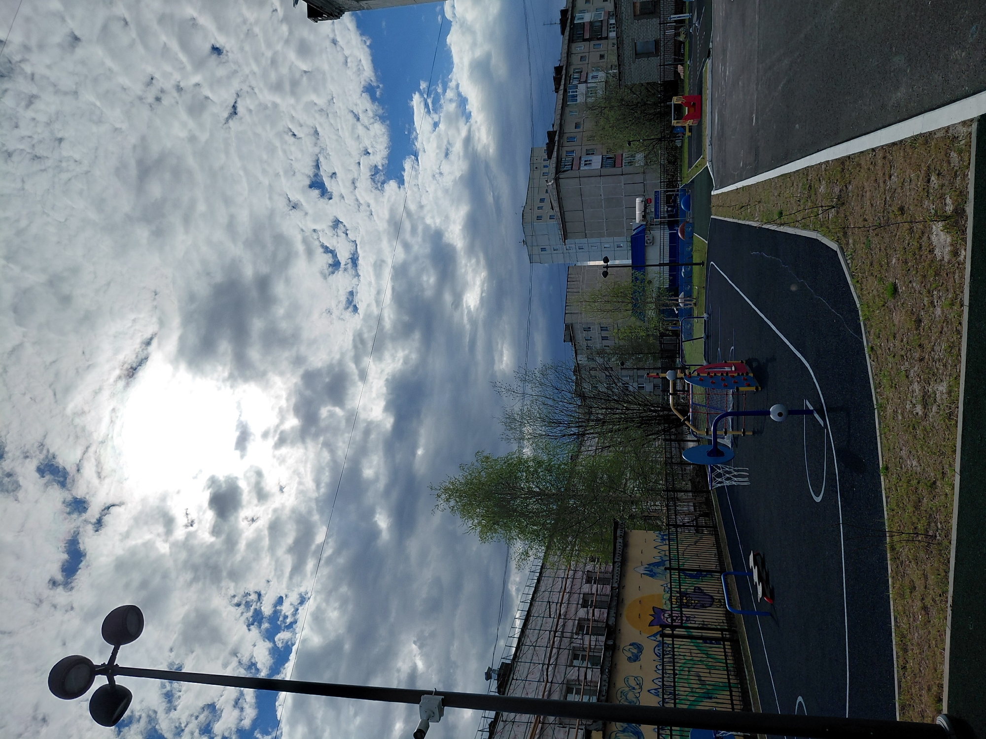 This photograph captures an urban outdoor scene in Nadym, Russia, featuring a residential area and a recreational space under a dynamic sky. The image appears to be taken during the daytime, with the sun brightly shining through breaks in a partly cloudy sky, creating a strong glare in the upper left.
The sky, occupying the upper and middle sections, displays a mix of white and grey cumulus clouds against patches of clear blue. In the mid-ground, several multi-story residential buildings, typical apartment blocks, line the horizon. Some of these buildings show signs of construction or renovation, with scaffolding visible on one structure. Trees with sparse, light green foliage suggest early spring or summer.
The foreground and lower-right depict a modern recreational area. This includes an asphalt-paved sports court marked with white lines, likely for basketball, as a hoop and backboard are clearly visible. Adjacent to the court, there is colorful playground equipment with blue and red components. A black metal fence separates areas, and a wall behind it features vibrant graffiti. Patches of dry grass and dirt border the paved surfaces. A prominent street lamp with three light fixtures on a pole stands in the lower left, extending towards the sky.
No people are visible, and no specific activity is taking place, but the presence of the court and playground suggests a space designed for community recreation.