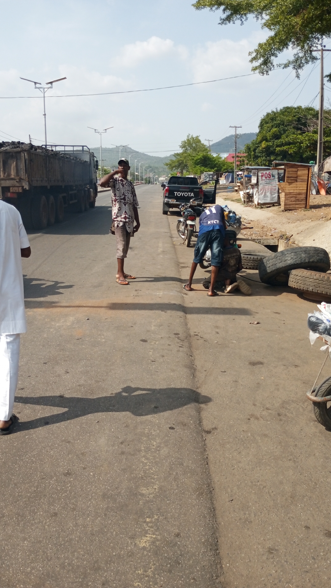 Location: Adankolo, Nigeria.
Scene: An open asphalt road in Adankolo. To the left, a large truck filled with dark materials, possibly coal, is partially visible. A man in a patterned shirt and cap stands on the road, his hand raised to his head, as if saluting. To his right, another man wearing a blue shirt with “KTCT” printed on it, and shorts, is bending down towards an object, perhaps working on it. A black Toyota pickup truck is parked in the middle of the road further ahead with a motorcycle parked nearby. In the background, there are other buildings, trees, hills, and utility poles. To the right, there is a small wooden structure, possibly a business or stall, with signage. Tires are on the street and there are other people in the scene.