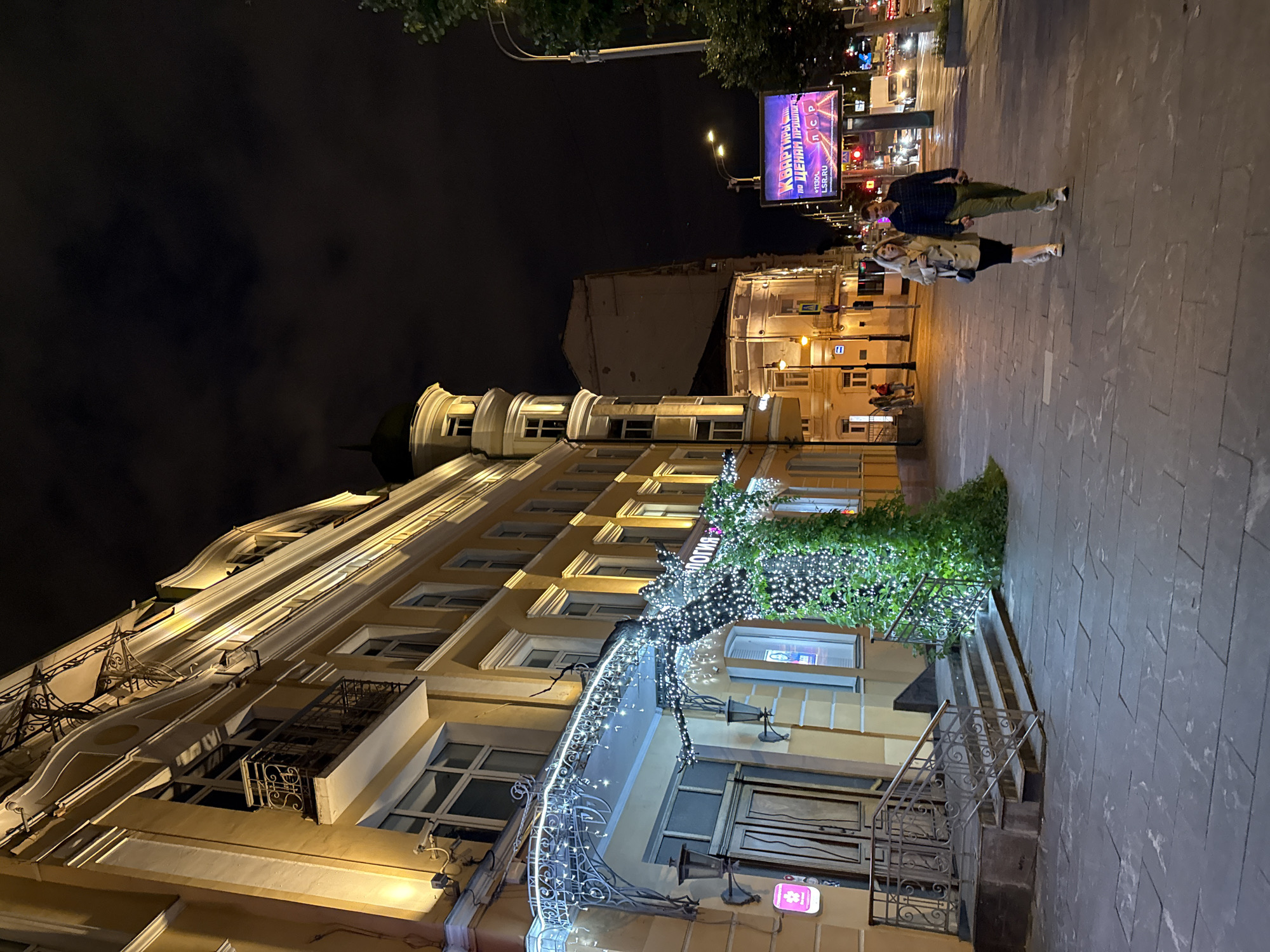 The image depicts a nighttime scene on a city street in Moscow, Russia. In the foreground, a couple walks hand-in-hand on a wide, paved sidewalk. The man wears a blue plaid shirt and olive green pants, while the woman is dressed in a tan trench coat and a black skirt. To their left, a building with a yellow facade is illuminated by warm lights. The building features ornate architectural details, including balconies with wrought-iron railings and decorative moldings. A festive display of white string lights adorns the entrance and a tree-like structure covered in greenery and lights. Stairs lead up to a recessed entrance with double doors. Further down the street, traffic lights are visible, indicating an intersection. A large digital billboard displays an advertisement in Russian. The sky is dark and cloudy, typical of a night scene in Moscow.