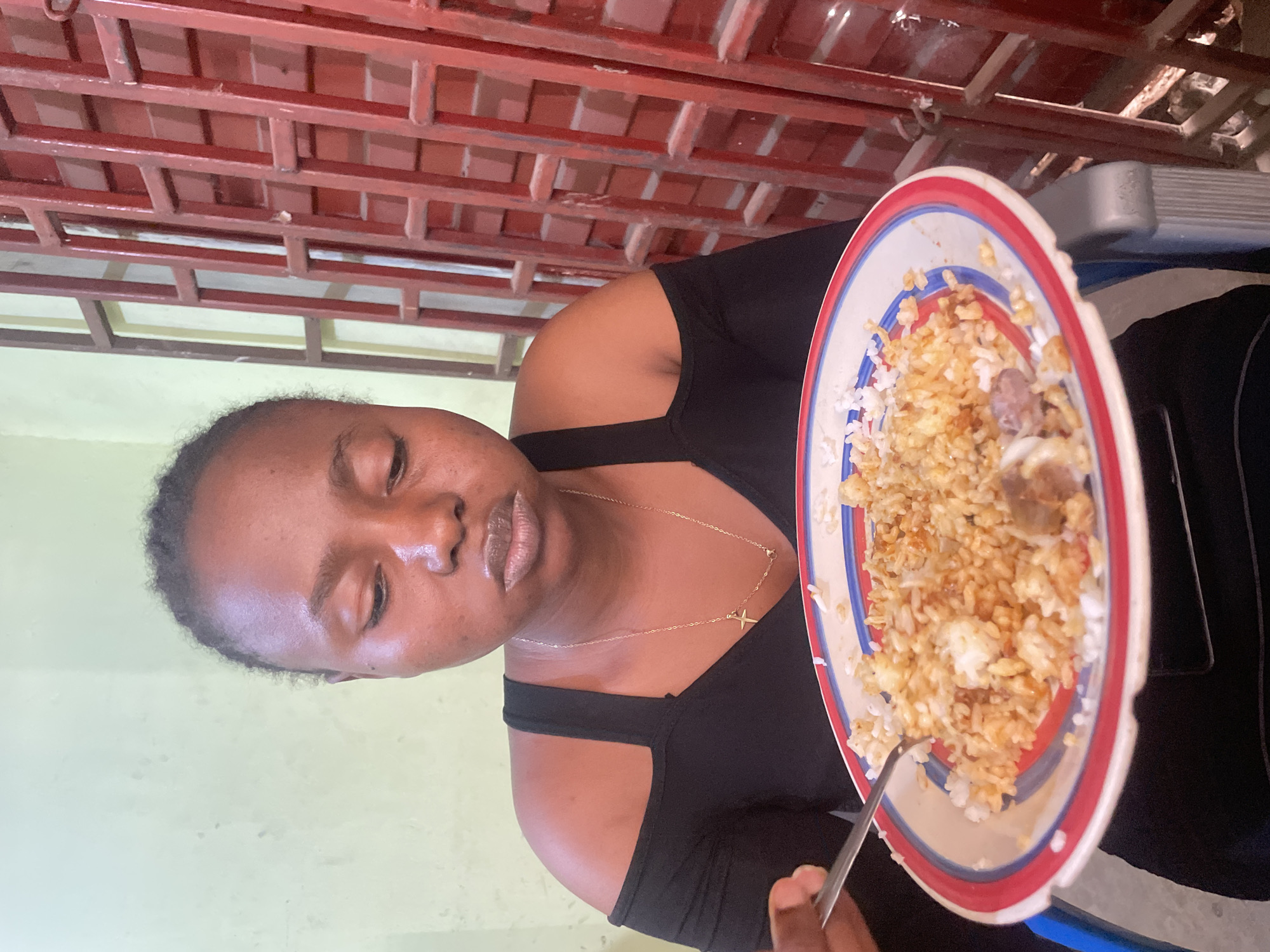 A person is eating a plate of rice with meat in an indoor setting. The person is wearing a black top and a gold necklace with a cross pendant. The environment includes a wall painted in a light yellowish hue and a red ceiling structure. There is a wheelchair armrest visible in the background. There is no verifiable information available in the image to confirm its location in the city of Jos, Nigeria.