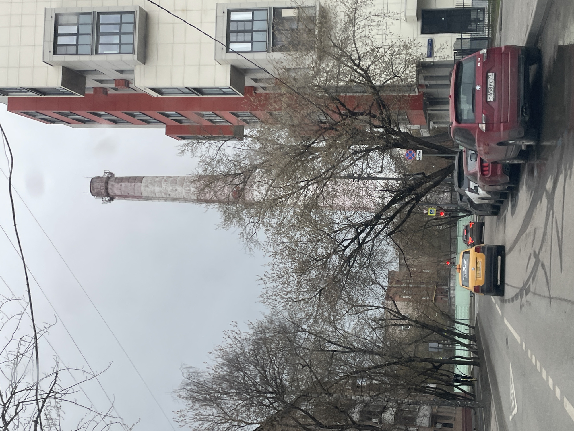 The image captures a street scene in an urban area on an overcast day. A tall, industrial chimney stands prominently in the background, constructed of red and white masonry. A modern apartment building with white and red facade sections occupies the left side of the frame. A row of bare trees lines the street, their branches obscuring parts of the buildings. Automobiles are visible on the right side of the street, including a red SUV with a license plate beginning with "0458 PC 77", a gray sedan, and a yellow taxi. The asphalt road shows signs of wear. Street signs and a traffic light are present.