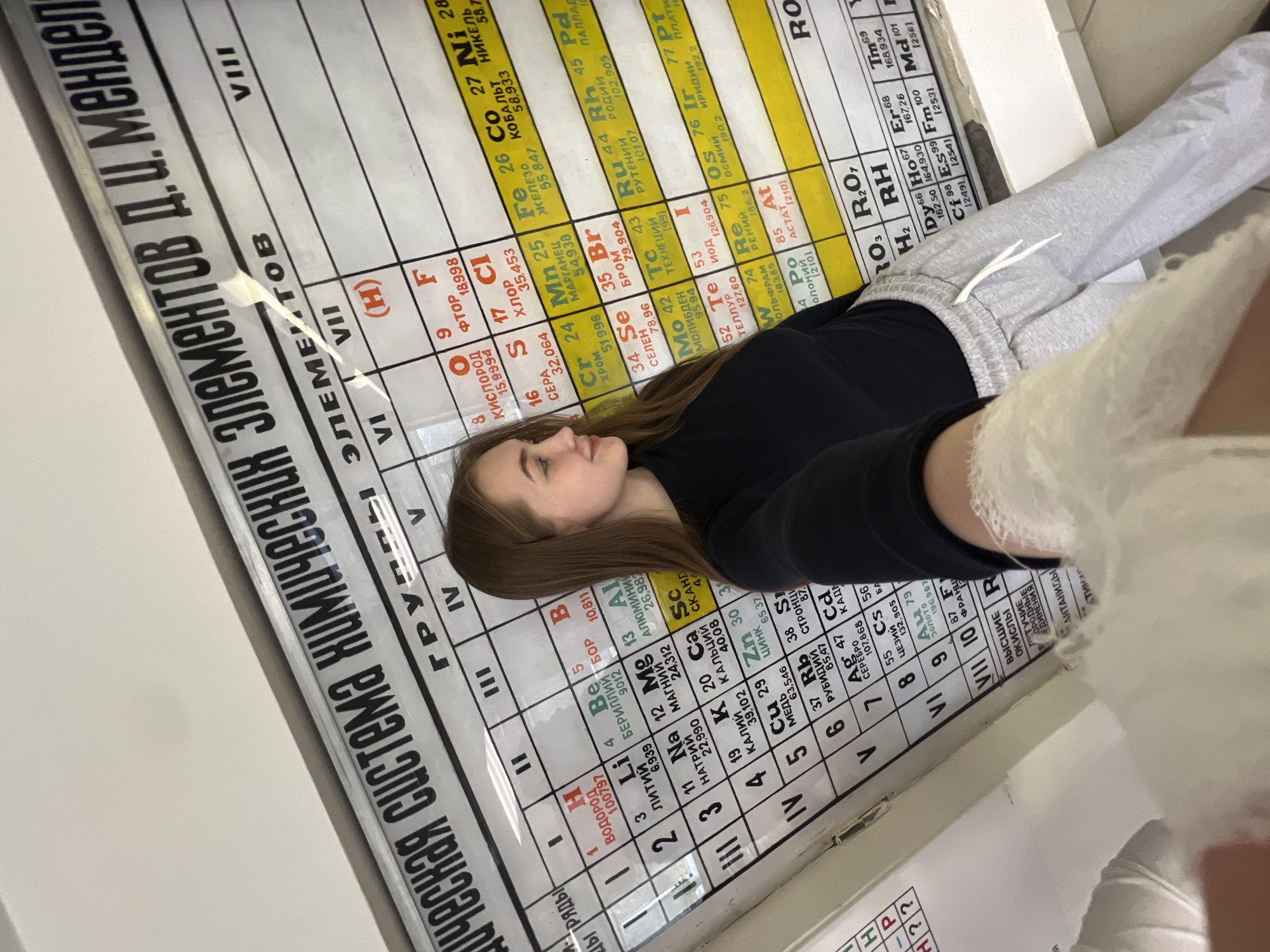 An indoor, medium shot depicts a young woman in Chaikovsky, Russia, resting or reclining against a large periodic table of chemical elements. She has long, straight brown hair and is dressed in a black long-sleeved top and light grey sweatpants with white drawstrings. Her right arm, closer to the viewer, is bandaged or in a white cast, suggesting a recent injury. Her gaze is directed upwards and slightly to her left, with a calm, neutral expression.
The prominent background is a large, laminated periodic table, likely a wall chart or poster, with text predominantly in Cyrillic. The main title at the top clearly reads "ПЕРИОДИЧЕСКАЯ СИСТЕМА ХИМИЧЕСКИХ ЭЛЕМЕНТОВ Д.И. МЕНДЕЛЕЕВА" (Periodic System of Chemical Elements D.I. Mendeleev). Visible element entries include "ВОДОРОД" (Hydrogen), "ЛИTИЙ" (Lithium), "КИСЛОРОД" (Oxygen), "ФТОР" (Fluorine), "ХЛОР" (Chlorine), "ЖЕЛЕЗО" (Iron), and "СЕЛЕН" (Selenium), among many others, all with their respective atomic numbers and masses. The chart also displays Roman numerals for groups (e.g., "VIII", "VII", "VI", "V", "IV", "III", "II", "I") and Arabic numerals for periods ("РЯДЫ"). The setting appears to be a classroom or a learning environment, illuminated by artificial indoor lighting.