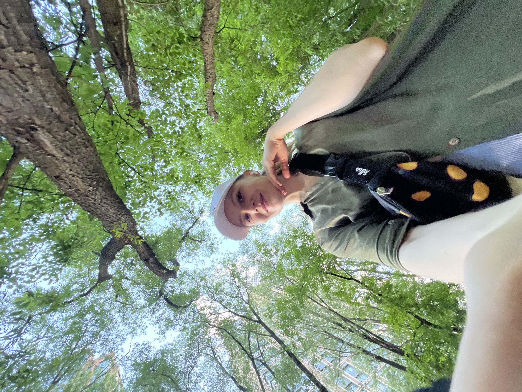 The image shows a person from a low-angle perspective. The person is wearing a white cap, a green shirt, and a black and yellow patterned bag. They are standing beneath a canopy of trees with green leaves. In the background, there are buildings visible between the trees.
The location of Khimki, Russia is not evident in the image.