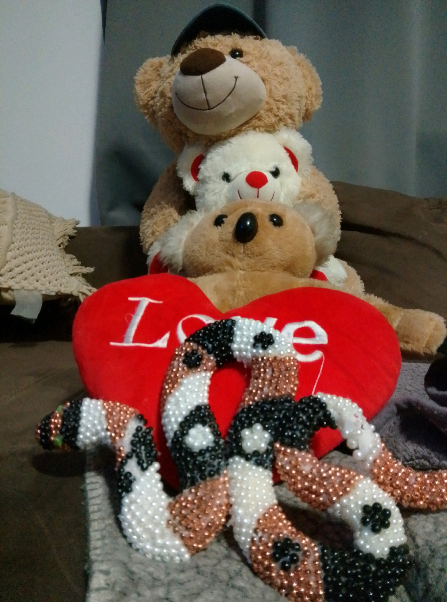 The image shows a collection of stuffed teddy bears and decorative candy canes arranged on a brown surface, likely a sofa. The largest teddy bear, at the top, is light brown and wears a dark blue baseball cap. Beneath it sits a smaller white teddy bear with a red nose and red accents on its ears, embraced by a medium-sized light brown teddy bear. In front of the teddy bears is a red heart-shaped pillow with the word "Love" embroidered in white. In the foreground, several candy canes, adorned with white, black, and copper-colored beads, are placed in front of the heart pillow. The background is blurred, showing a textured, possibly grey, wall. The scene appears to be indoors, in a domestic setting. There is no identifiable location in the city of Wedel, Germany.