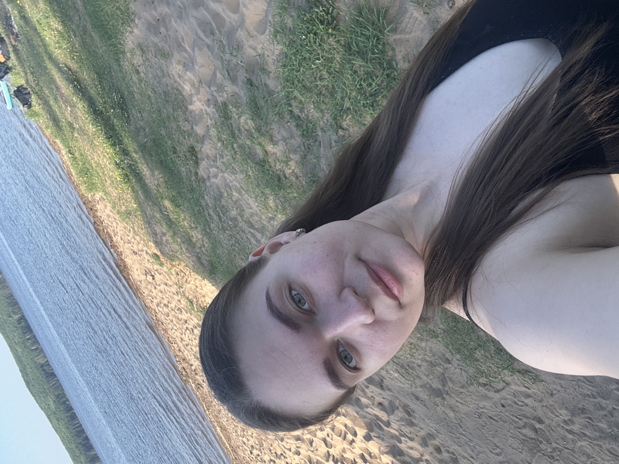 A young woman is taking a selfie on a sandy beach. She has long brown hair and is wearing a black top. Behind her is a body of water and a grassy area. The location appears to be the shoreline of the Kama River near the city of Chaikovsky, Perm Krai, Russia, based on the typical environment of the region. There is a small boat parked near the grass area.