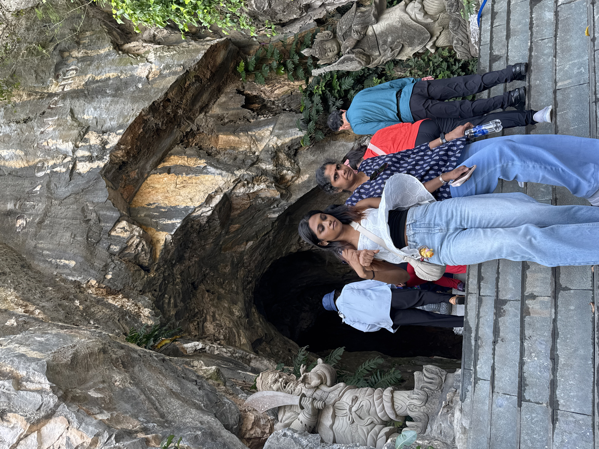 The image displays the entrance to Động Âm Phủ (Hell Cave) at the Marble Mountains (Ngũ Hành Sơn) in Đà Nẵng, Vietnam. A large, dark, irregularly shaped cave mouth is recessed into a massive, textured grey and brown rock face. The text "ĐỘNG ÂM PHỦ" is carved into the rock above the entrance. Two intricately carved stone guardian statues flank the cave opening: a figure on the left brandishing a large, ornate sword, and a figure on the right in armor holding a musical instrument or scepter. Wide, grey stone steps descend from the cave entrance towards the foreground.
In the immediate foreground, two women are present. The left-most woman, with dark hair and light complexion, wears a white strapless top, light blue wide-leg jeans, and a light-colored crossbody bag with a yellow charm attached; she looks downwards to her right. Adjacent to her, an older woman with dark hair, wearing a patterned dark blue short-sleeved top and light blue wide-leg pants, holds a clear plastic water bottle in her right hand and a light-colored smartphone in her left, looking towards the viewer.
In the midground, several individuals are positioned near the cave entrance. A person in a light blue long-sleeved shirt, dark pants, and a blue bucket hat faces the cave entrance, ascending the steps. Partially obscured by this person, another individual in a red long-sleeved top and dark pants also faces the entrance. To their right, a man in a teal long-sleeved shirt and dark pants stands facing the cave entrance area. Sparse green vegetation grows from crevices in the rock faces, particularly on the right side. The scene is illuminated by natural daylight.