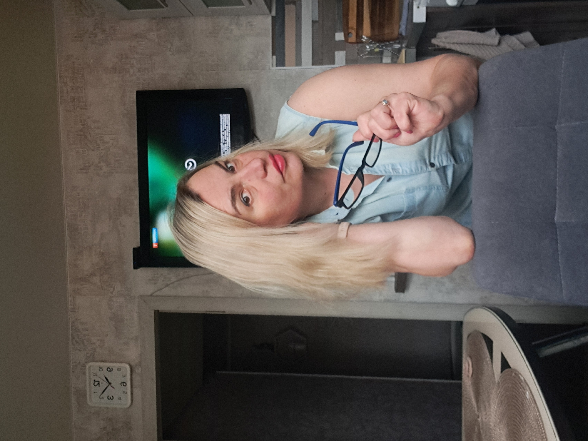 A woman with blonde hair is holding a pair of blue-framed glasses. She is wearing a light blue shirt. Behind her, there is a television and a wall-mounted clock that reads approximately 3:15. The setting appears to be indoors, possibly in a home in Elektrostal, Russia, although there are no elements in the photo to confirm the location.