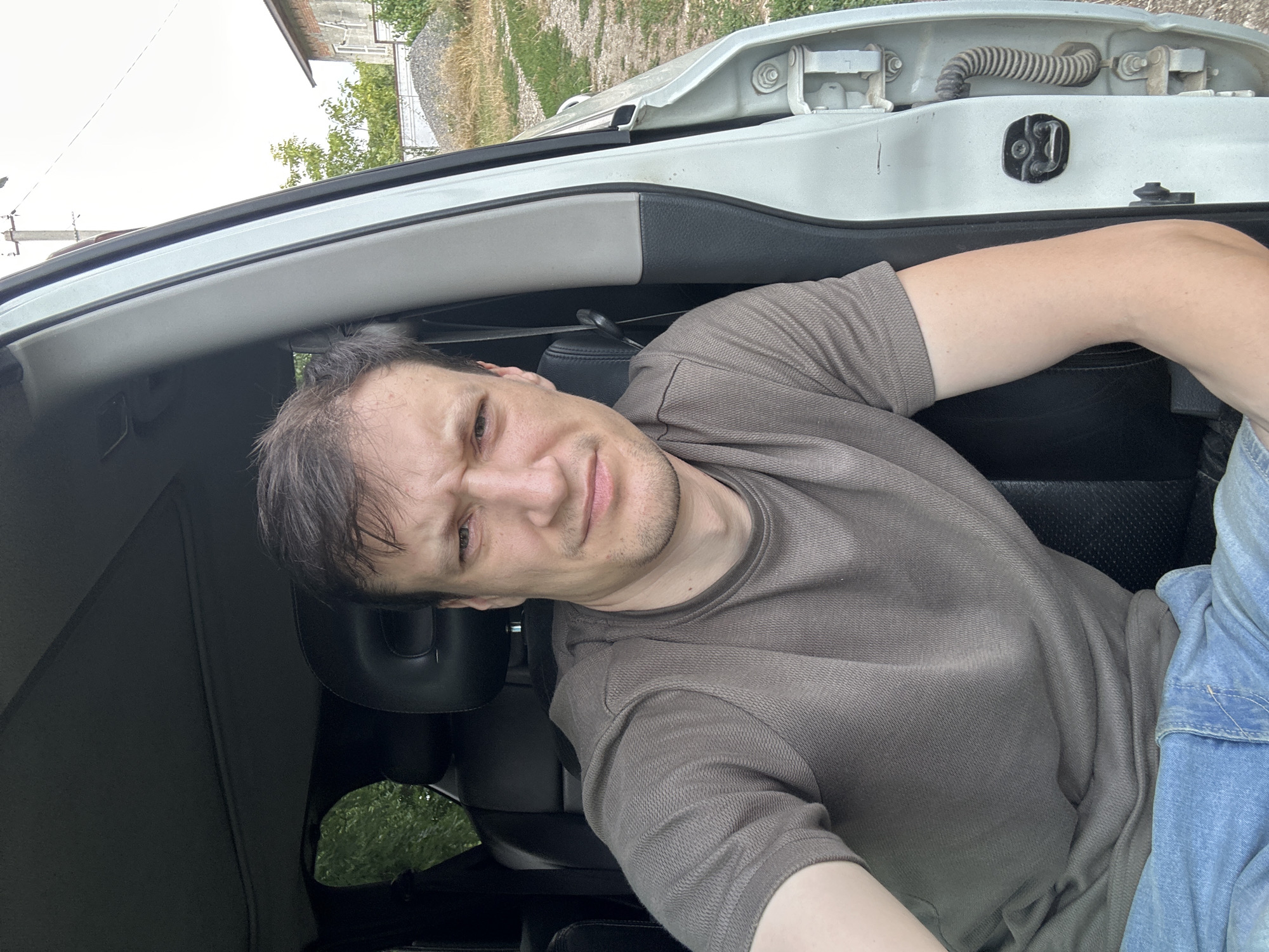 A medium shot shows a man with dark hair and a neutral expression, sitting in the driver's seat of a white car with the door open. He is wearing a brown t-shirt and blue jeans. The car interior, including black leather seats, is visible. The exterior of the car shows a portion of the door and frame, with some rust on the hinges. Behind the car, a dirt road and overgrown grass are visible. In the background, a small brick building and some trees suggest a rural or suburban setting. The sky is overcast. The image appears to be a selfie taken by the man. Based on the user's request, the image is situated in Makiv, Ukraine.