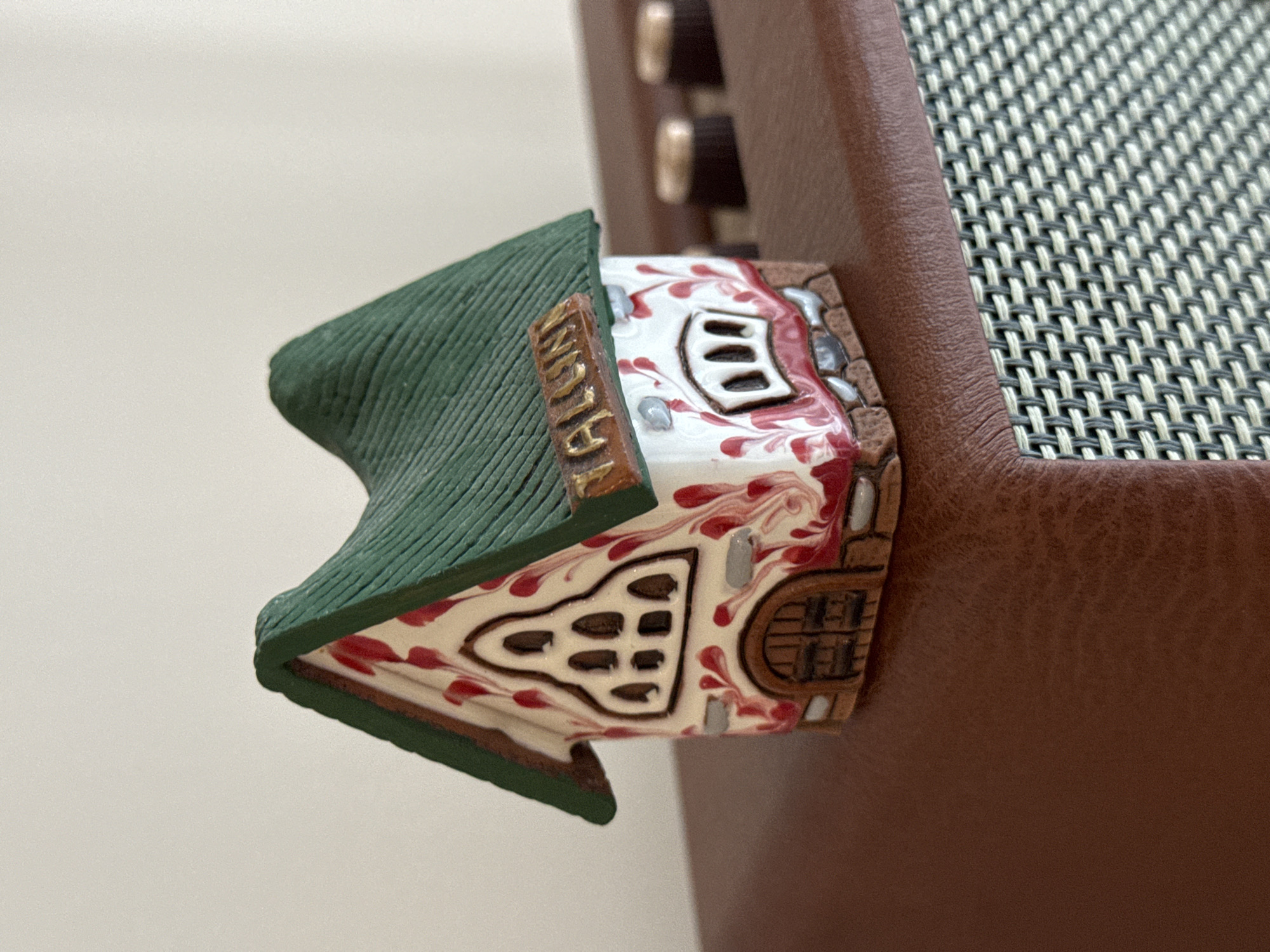 A miniature, white house with red decorative streaks and a green roof is placed on top of a brown leather-covered object. The house has a sign on its roof that reads "YALIN." The brown object appears to be a piece of electronic equipment, possibly an amplifier, with visible knobs and a textured grill. The background is a plain, light-colored wall. The scene is indoors, with artificial lighting. There are no people or animals present, and no specific location in Izhevsk, Russia, can be identified from the image.