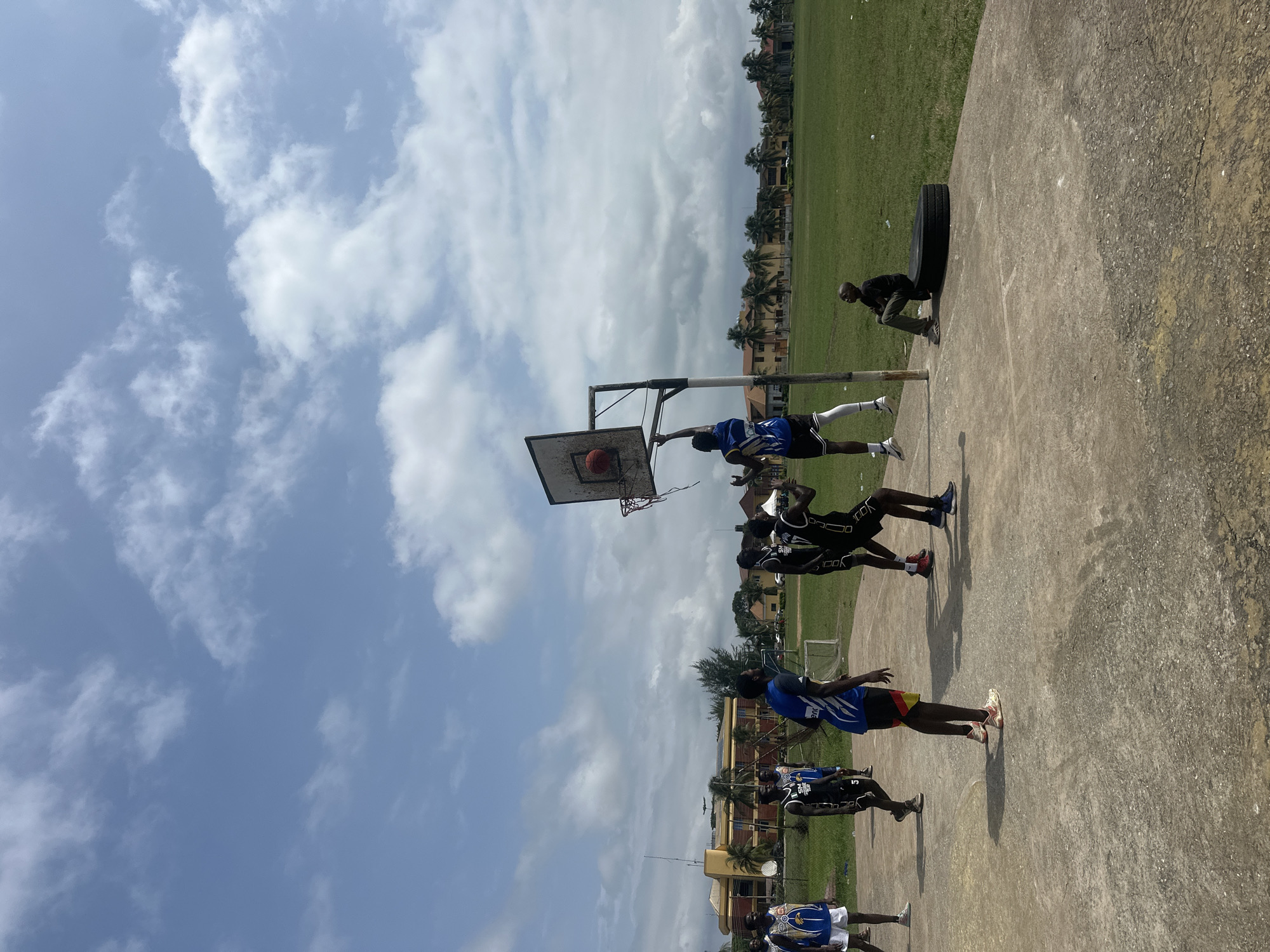 The image captures a basketball game in progress on an outdoor court in Ajara, Nigeria. Several young men, dressed in basketball attire, are actively engaged in the game, with one player in mid-air attempting a shot at the hoop. Another individual is seated on a tire at the side of the court, observing the game. The court is made of concrete, with some visible cracks and wear. In the background, there are buildings, palm trees, and a vast expanse of green grass, suggesting a semi-urban or tropical environment. The sky is partly cloudy, with bright sunlight indicating daytime. The scene depicts a typical recreational activity taking place within a community setting.