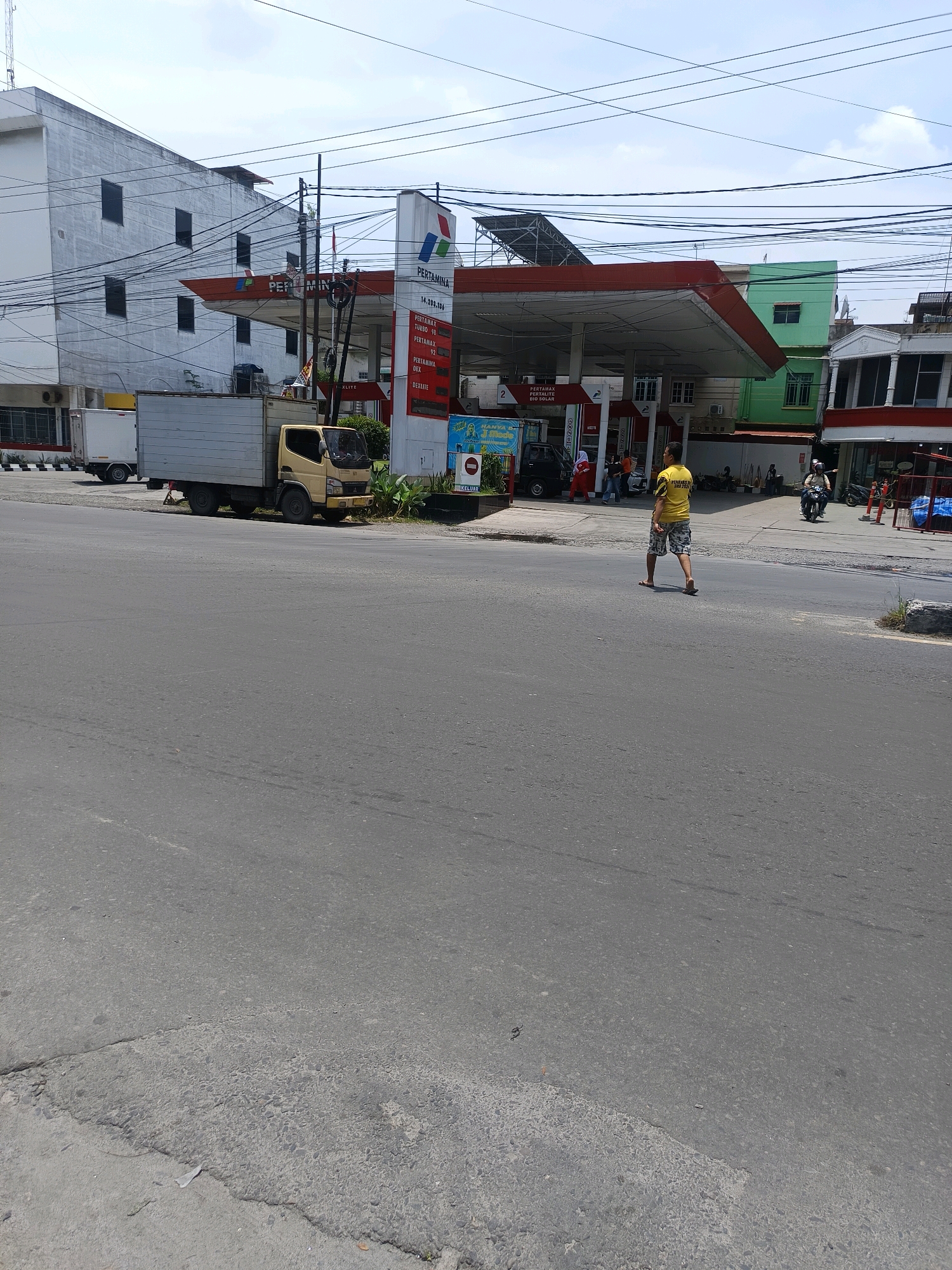 The image shows a street scene in Percut Sei Tuan, Indonesia, featuring a Pertamina gas station. A man in a yellow shirt and patterned shorts is walking across the street away from the gas station. To the left, a light-colored truck with a grey cargo box is parked, and behind it, a white truck is partially visible. The Pertamina gas station has a red roof supported by white columns, with fuel pumps under the canopy. Signs indicate fuel prices, including "PERTAMAX TURBO 98," "PERTAMAX 92," "PERTAMINA DEX," and "BIO SOLAR." Multiple buildings are visible in the background, including a white multi-story building on the left and a green building to the right of the gas station. Power lines crisscross the sky. A motorcycle with a rider is seen in the middle ground near the gas station. The road surface appears to be asphalt with some cracks and patches. The sky is partly cloudy.