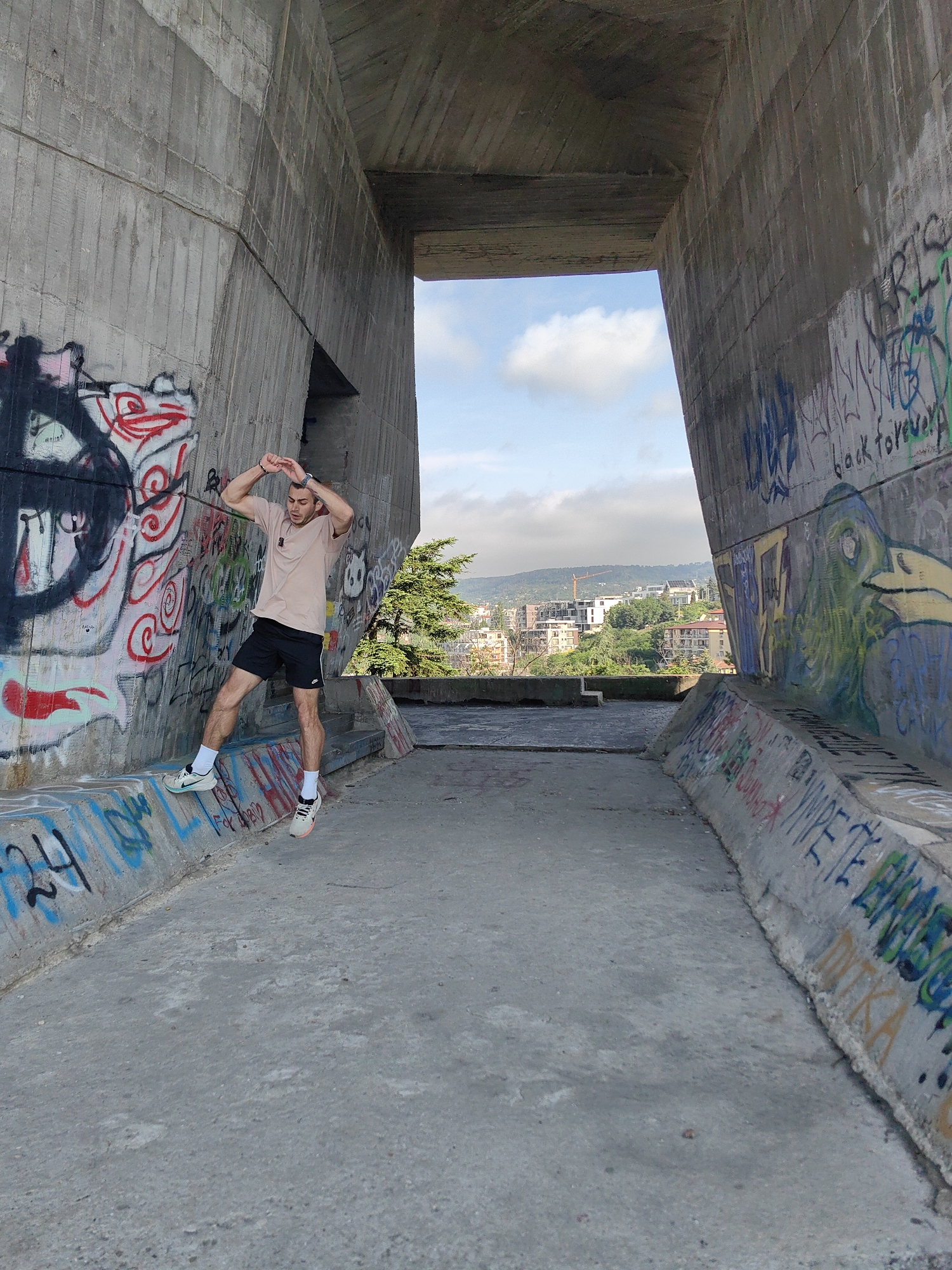A young man stands with his arms raised, hands touching his head, in an open, concrete structure under a bridge or overpass. He is wearing a light tan t-shirt, black shorts, white socks, and athletic shoes. The concrete walls are heavily graffitied with colorful spray paint. The ground is also concrete. In the background, through the opening of the structure, a city landscape with buildings and trees is visible under a cloudy sky. The scene is likely located near the Varna City Mall, as this structure is a known landmark in that area, possibly part of the surrounding infrastructure.