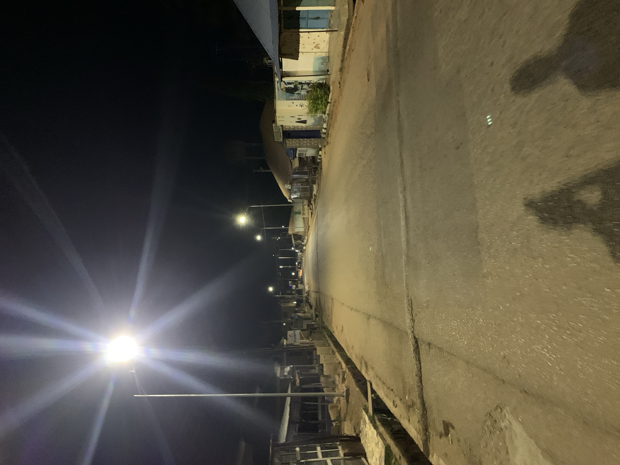 An eye-level view of a street in Kabong, Nigeria at night. The street is paved and bordered on both sides by buildings, mostly low-rise structures. Streetlights illuminate the scene, casting long shadows and creating stark contrast. The road appears damp, reflecting some of the artificial light. There is no visible activity in the image, and no people or vehicles are present. The environment is urban, but appears quiet and deserted.