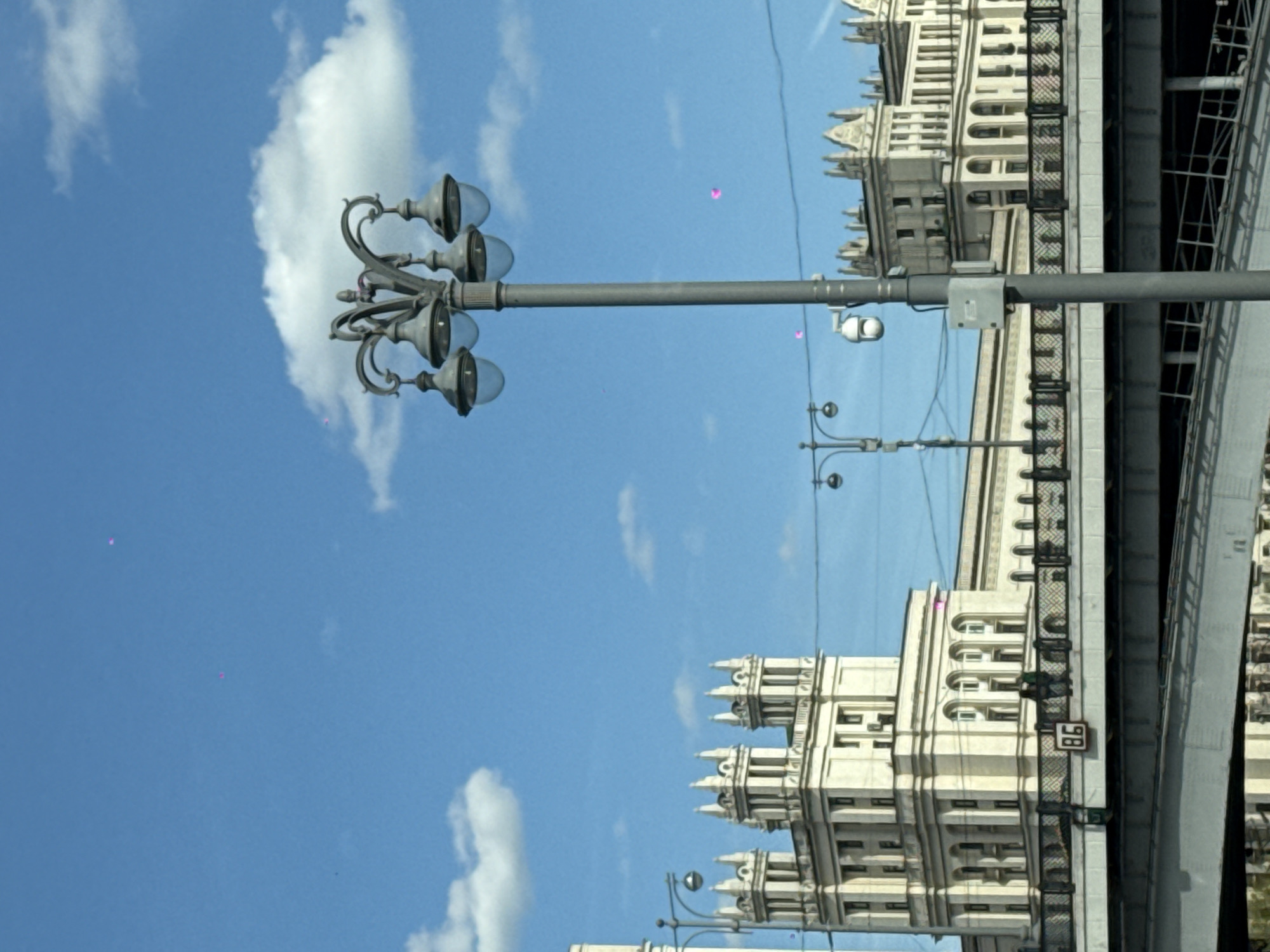 The image shows a view in Moscow, Russia, featuring a street lamp in the foreground. In the background, there are large, ornate buildings that are reminiscent of Soviet architecture. A bridge is situated next to the building. The sky is blue with white clouds. Other street lamps are also evident.