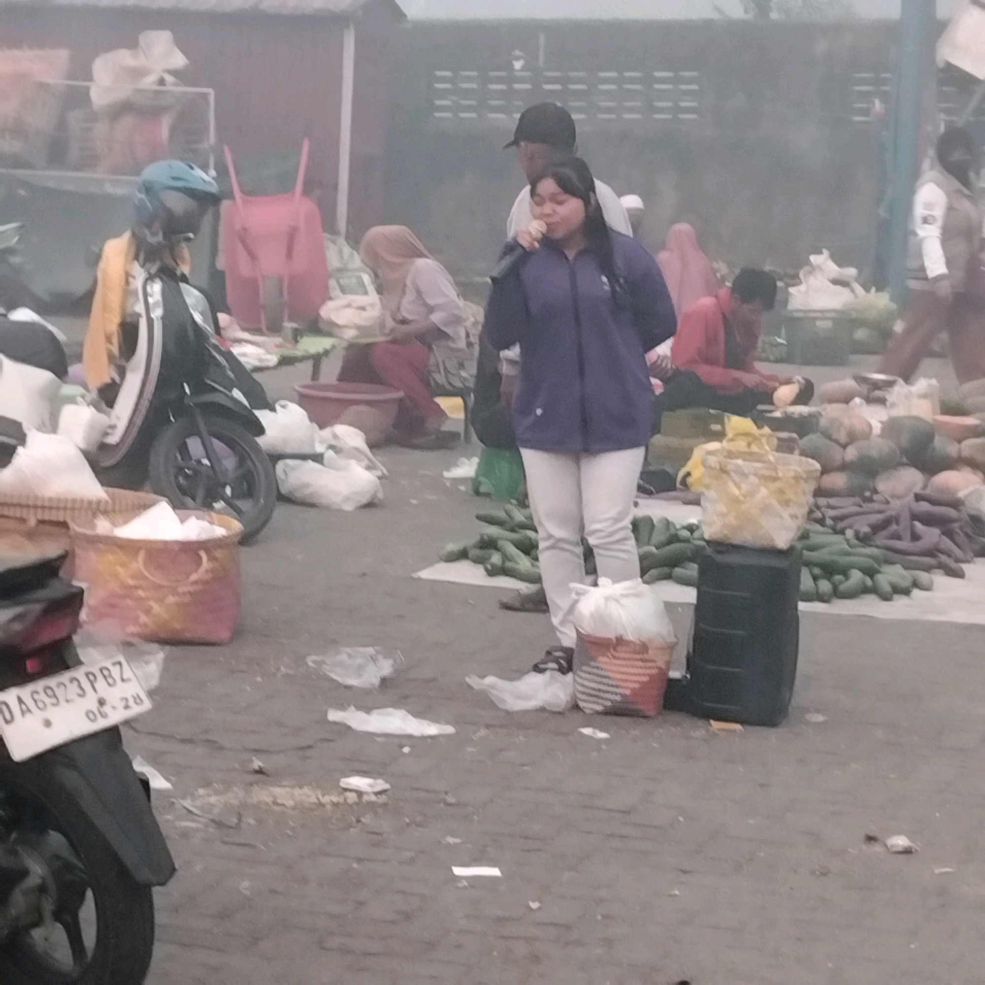 The image depicts an outdoor market scene, possibly in Banjarbaru, Indonesia, characterized by its hazy, overcast atmosphere. In the foreground, a woman in a purple jacket and light-colored pants is holding a microphone, appearing to address an audience or make an announcement. A man wearing a black cap stands behind her, with his arm around her shoulder. Scattered around them are various market goods, including vegetables like cucumbers and eggplants, as well as baskets, bags, and a motorcycle. Several individuals are visible in the background, some engaged in activities related to the market, such as selling or preparing produce. The ground is paved, and there is visible litter, suggesting a busy and somewhat unkempt environment. The overall impression is that of a typical, bustling local market.