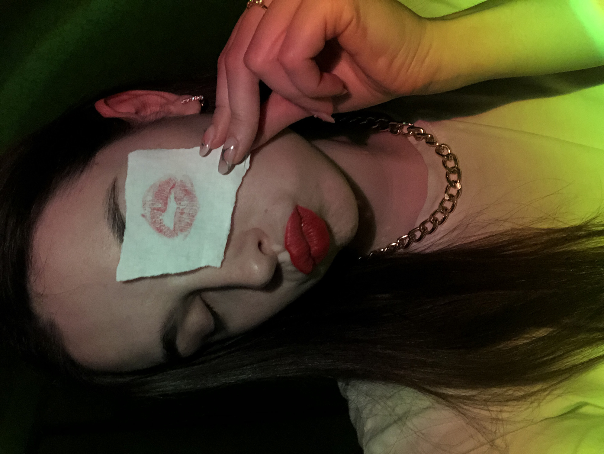 A woman with long, dark hair is lying down. A square of white paper with a red lipstick kiss is covering her left eye. She has red lipstick on her lips and is wearing a gold chain necklace. Her left hand is near her face, with manicured nails. She is wearing a ring on her left hand. The background is dark with some green and yellow lighting. The location is not specified, and there is no indication that the picture was taken in Noviy, Russia.