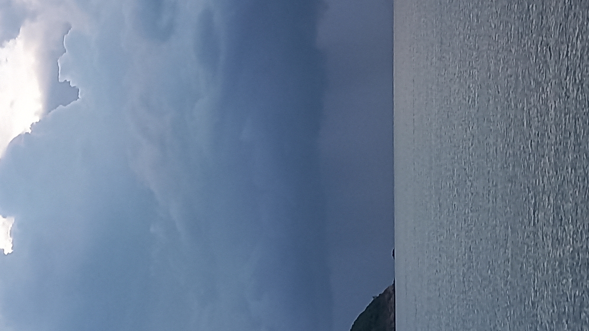 The image depicts a wide expanse of the sea under a dramatic, overcast sky. Dark, heavy clouds dominate the upper portion of the frame, with lighter, more diffused clouds visible near the top edge. A sliver of land, appearing as a green, tree-covered island or coastline, is visible on the far left, jutting into the sea. The sea itself is a muted grey-blue, with subtle ripples suggesting a calm to moderate swell. The horizon line is sharp and distinct, dividing the sea from the sky. The scene conveys a sense of impending weather change, possibly rain, due to the dark cloud formations. There are no people or man-made objects clearly visible in the image, emphasizing the natural, elemental quality of the landscape. The location is the Ko Pha-ngan District in Thailand, a region known for its tropical islands and coastlines.