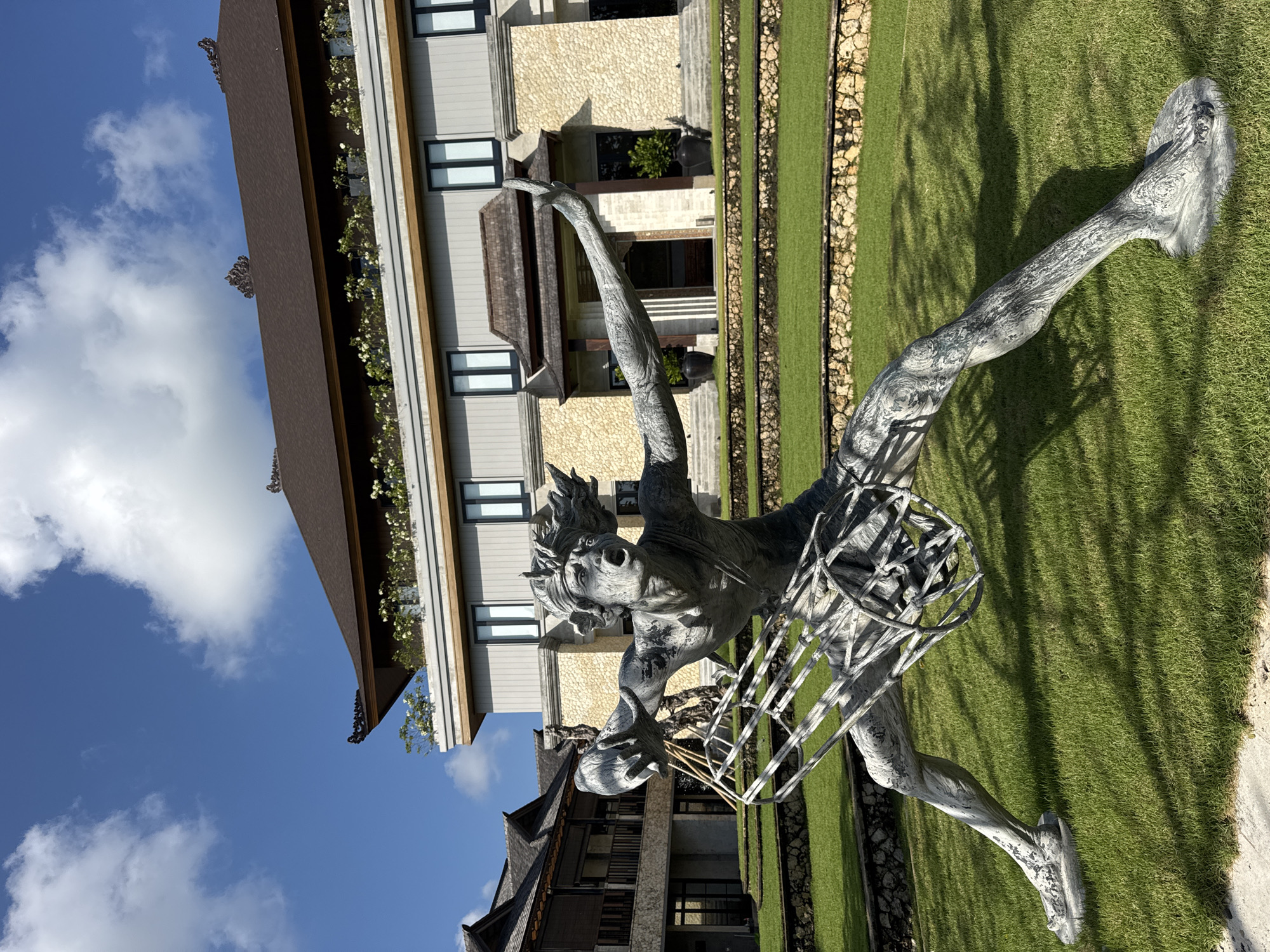 A gray, weathered stone sculpture of a human figure is positioned in the foreground on a meticulously manicured lawn. The figure is depicted in a dynamic, wide-legged stance, with its mouth open in a shout or gasp, and arms outstretched. The sculpture appears to be holding a cage-like structure. Behind the sculpture, a building with a traditional Balinese-style dark brown roof is visible. The building has multiple stories with light-colored stone and siding, and features several dark-paned windows. White flowers are planted in planters along the upper level of the building. To the left, another building with a similar architectural style is partially visible. The scene is set under a bright blue sky with scattered white clouds, suggesting a sunny day. The grass is well-maintained and bordered by stone walls. The location is identified as being in Kuta Selatan, Indonesia, likely at a resort or hotel given the architecture and landscaping.