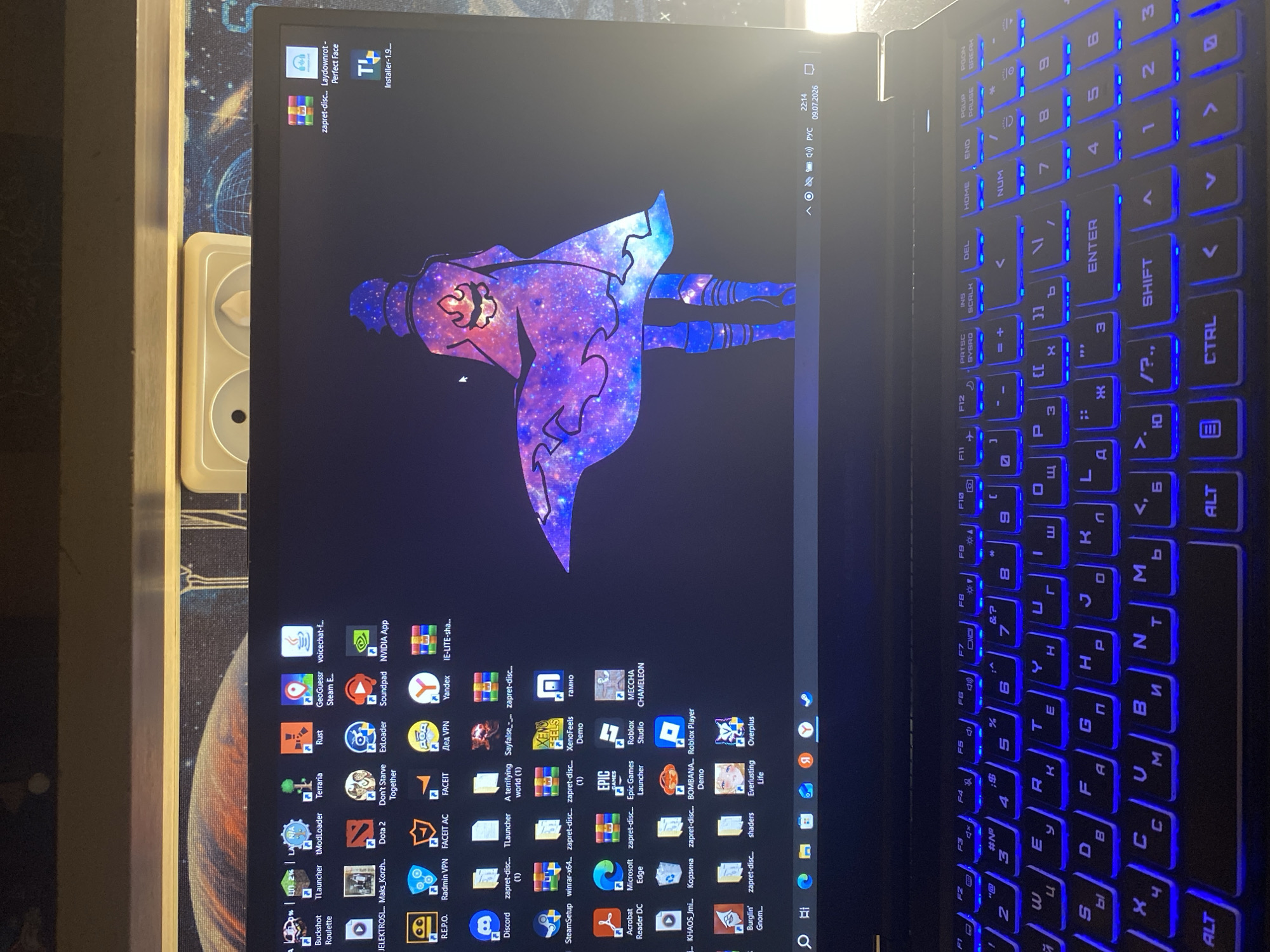 The image shows a close-up view of a laptop with a dark background on the screen. The screen displays a desktop with numerous application icons and a wallpaper featuring a stylized character dressed in a purple and blue galaxy-themed outfit. The character is depicted in a dynamic pose, seemingly floating or flying.
On the right side of the laptop, the keyboard is illuminated with vibrant blue backlighting. The keys are arranged in a standard QWERTY layout, with some Cyrillic characters also visible, indicating the potential for a Russian language setting. The keyboard is the primary focus of the right half of the image, showcasing the glowing keys.
The bottom right corner of the screen displays system information, including the time "22:14" and the date "09.07.2026". The presence of "PYC" suggests the Russian language is in use.
The scene is indoors, likely a room in a home or office, given the presence of the laptop and the ambient lighting. The overall mood appears casual, perhaps indicating the user is engaged in gaming or other computer-related activities. The distinct wallpaper suggests a personal preference for fantasy or anime aesthetics. There are no visible people in the image.