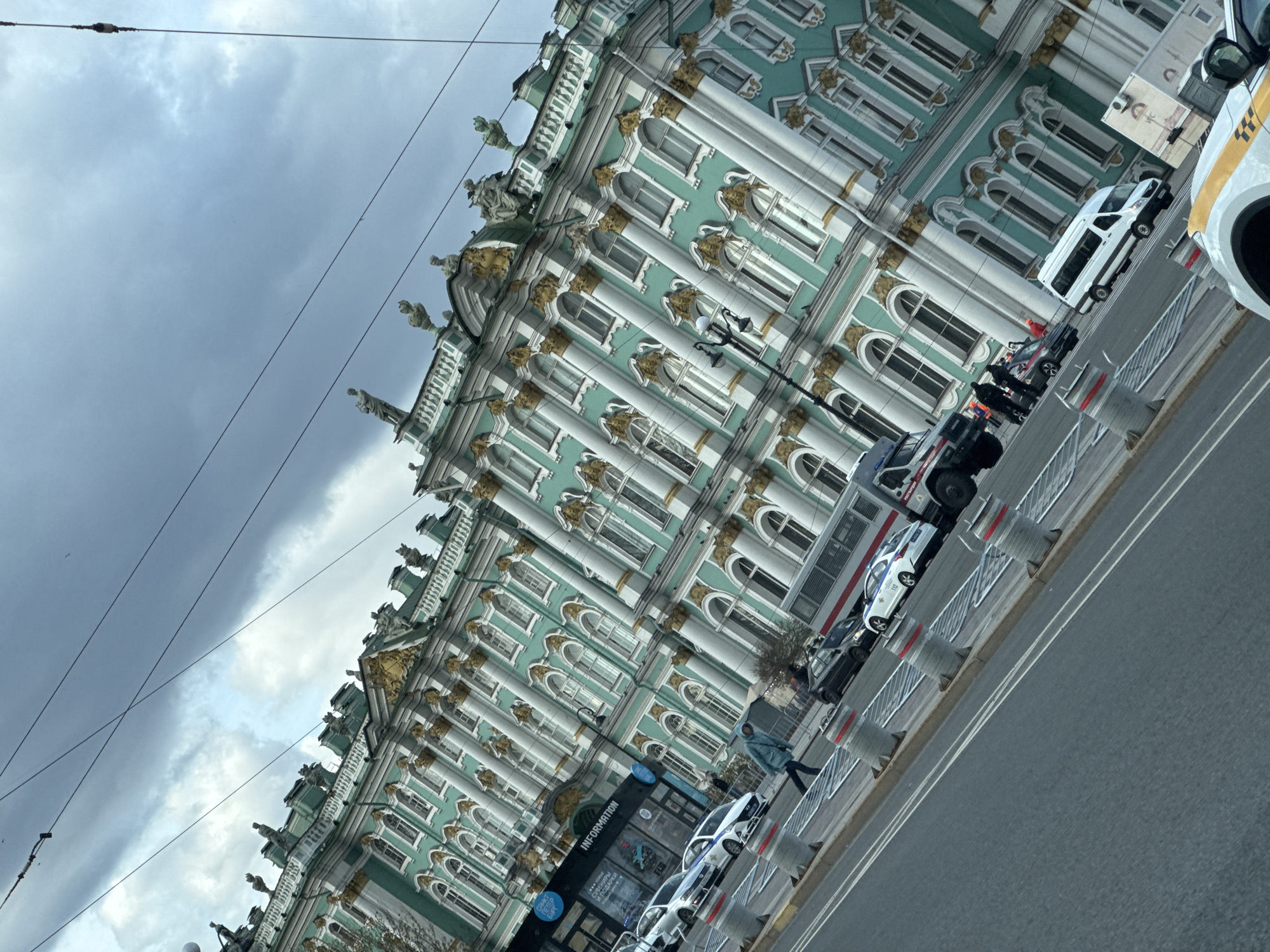 The image depicts a view of the Winter Palace in Saint Petersburg, Russia, under a cloudy sky. The palace's facade is adorned with columns, ornate decorations, and sculptures. Vehicles, including cars and a truck, are parked along the road in front of the building. A person is walking on the sidewalk near a sign that says "INFORMATION." The street is bordered by temporary barriers.