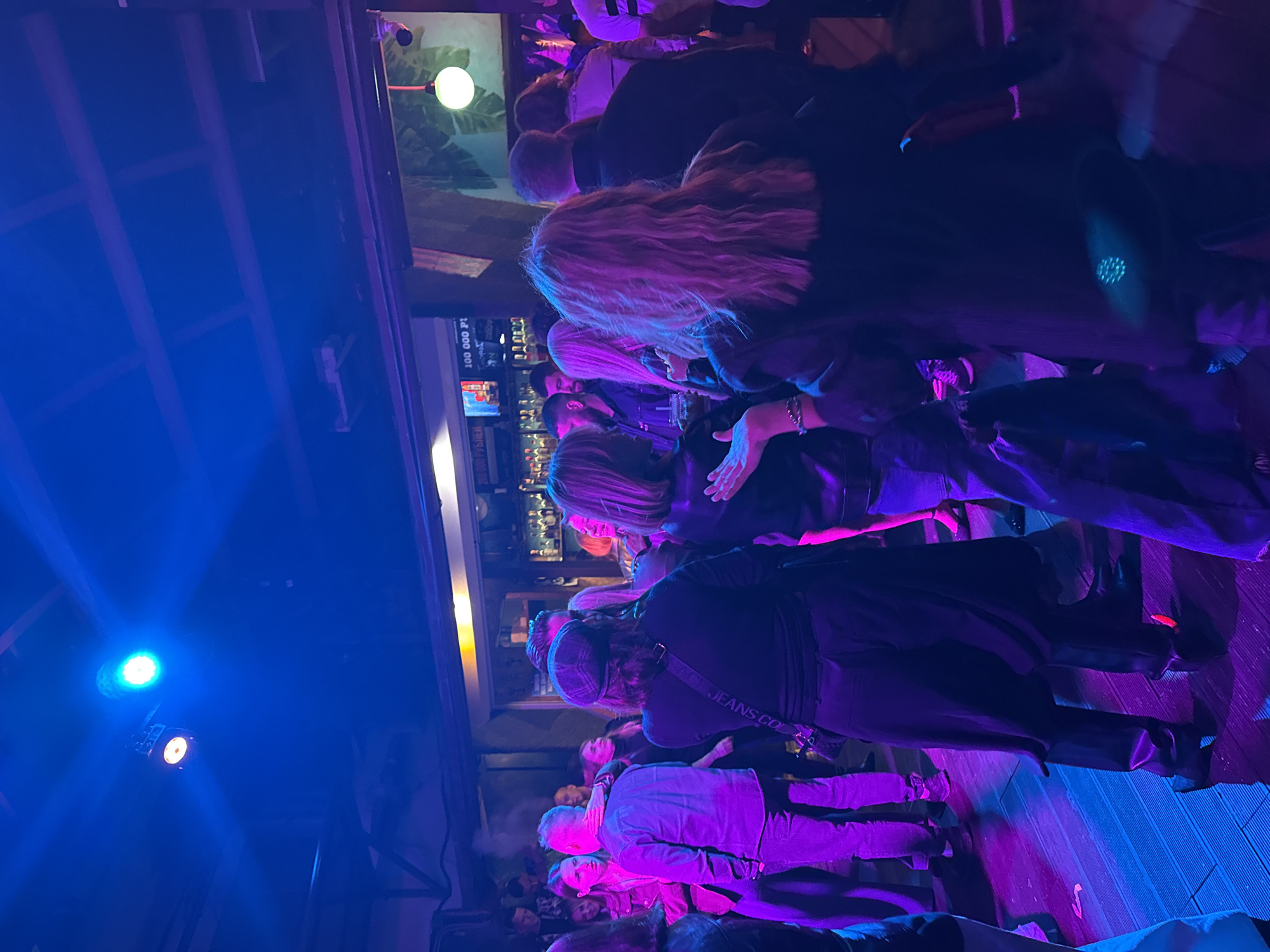A crowded indoor space, possibly a bar or nightclub, is illuminated by vibrant blue and pink lights. The scene depicts a gathering of people, some dancing and interacting. In the background, a bar with bottles is visible, and on the wall, a screen displays the text "100 000 P!". The floor appears to be wooden planks. The atmosphere suggests a lively social event in Perm, Russia.
